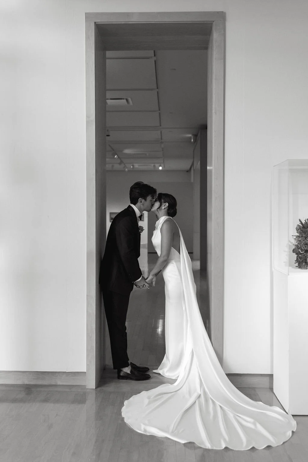 A black and white photograph of a wedding couple standing in a doorway, about to kiss, holding hands. The bride is wearing a long, elegant satin wedding gown with a train, and the groom is in a dark suit.