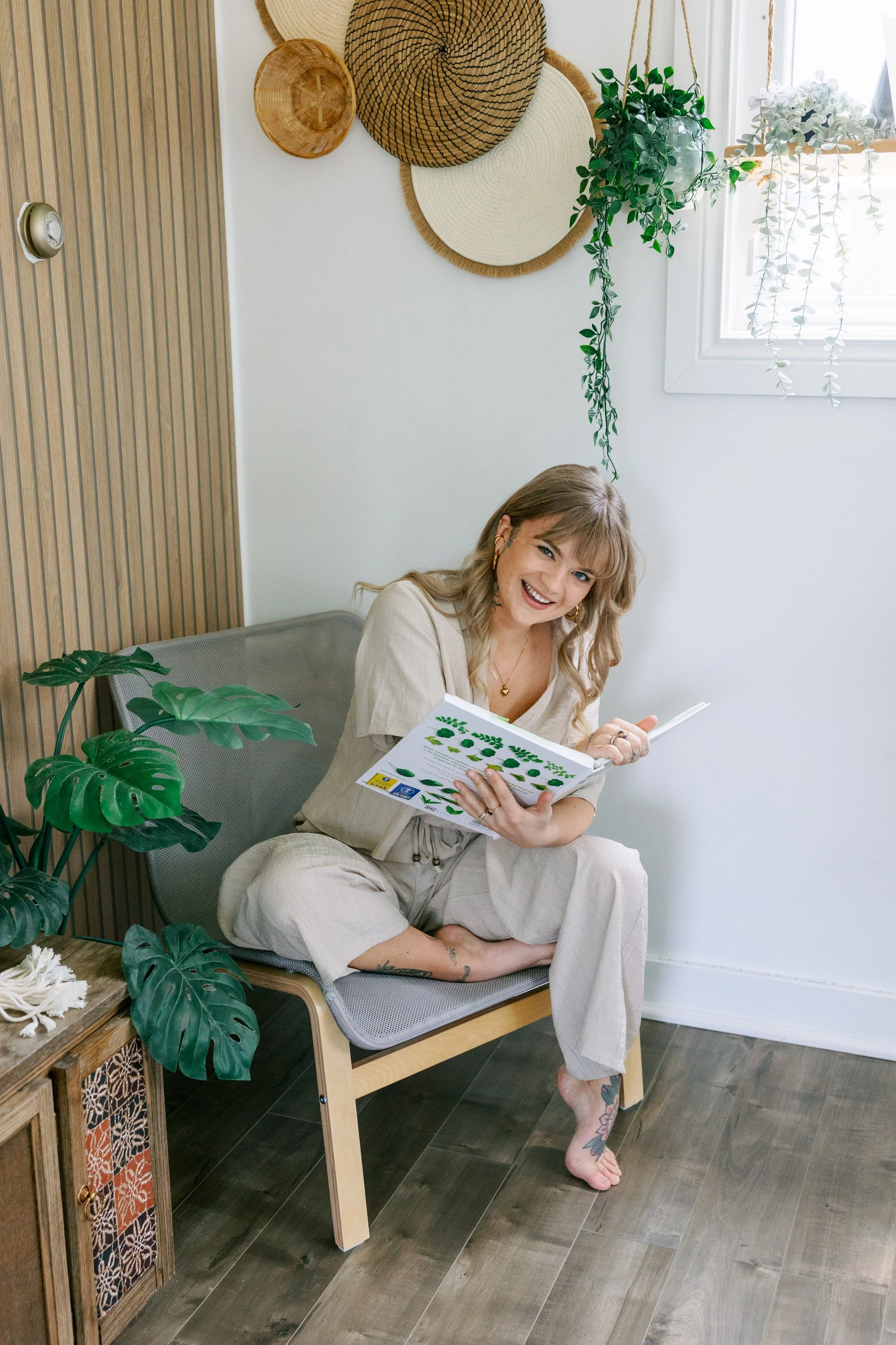 A woman sitting on a chair with a book, smiling. She has blonde hair, tattoos on her foot, and is barefoot. The room is decorated with hanging baskets, plants, and rustic furniture, with a window letting in natural light.