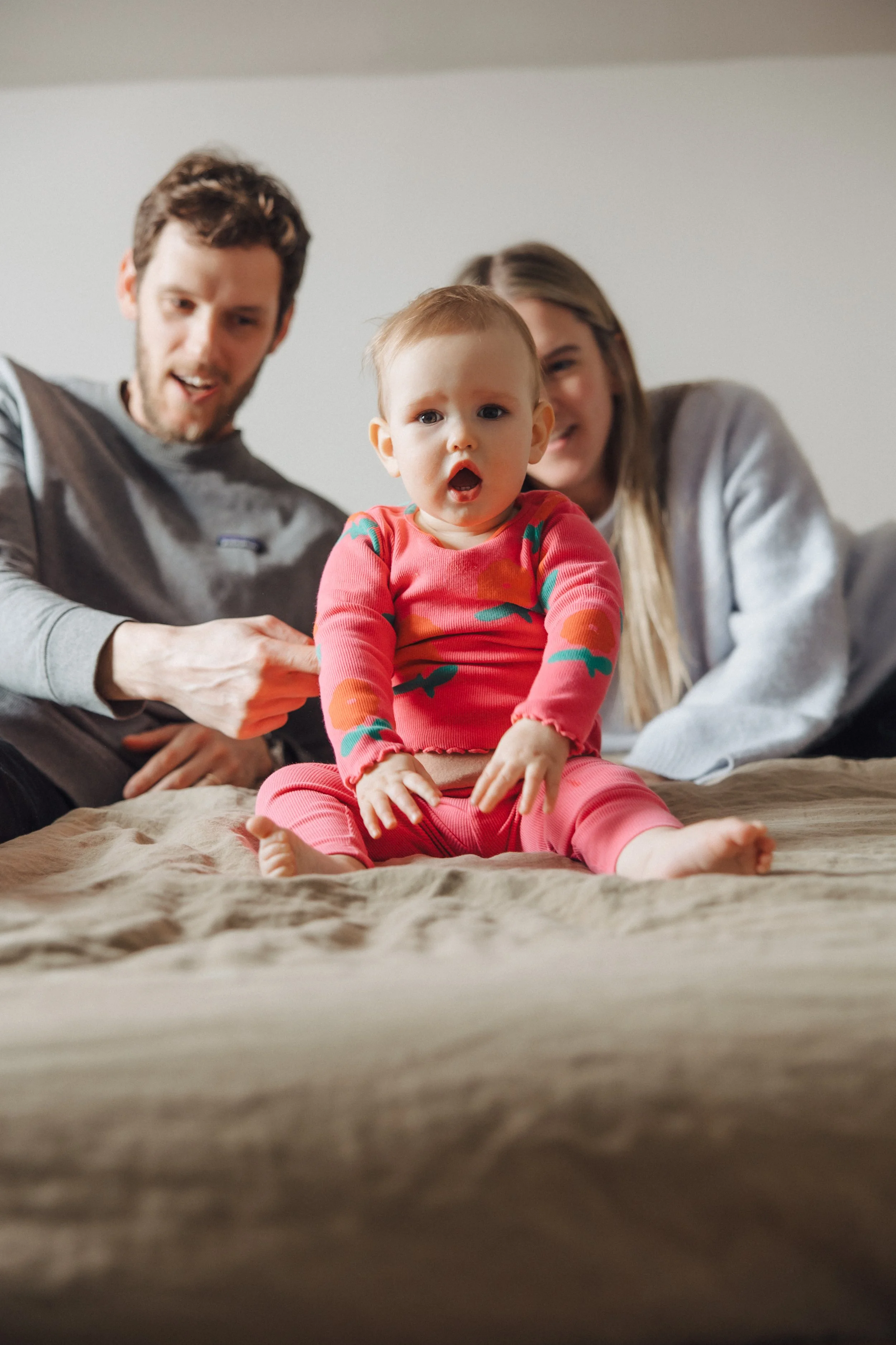 A young family, including a baby in a red and green patterned outfit, is sitting on a bed with a beige blanket. The baby looks surprised or curious, while the parents smile and look at the baby. The background is a plain wall.