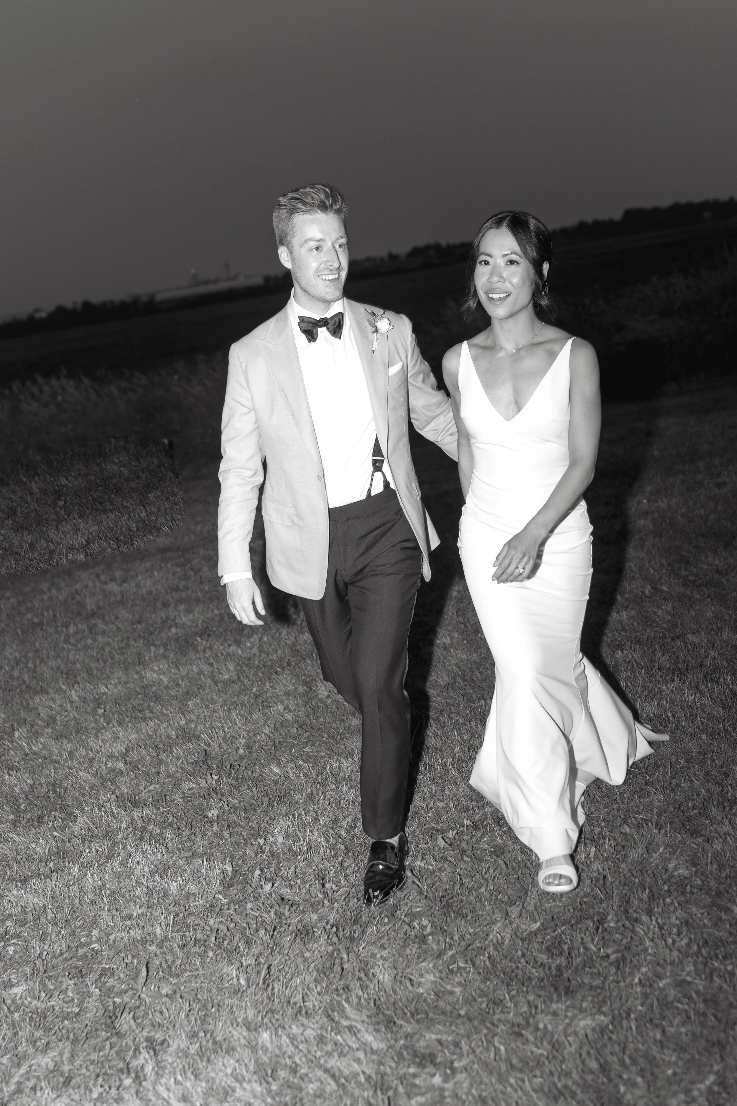 A black-and-white photo of a smiling couple walking outdoors at dusk or night, dressed in formal wedding attire, with the man in a light-colored suit and bow tie and the woman in a white sleeveless dress.