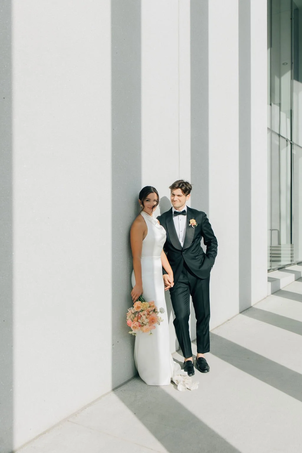 A bride and groom stand against a modern white building with large windows, holding hands and smiling softly, dressed in wedding attire.