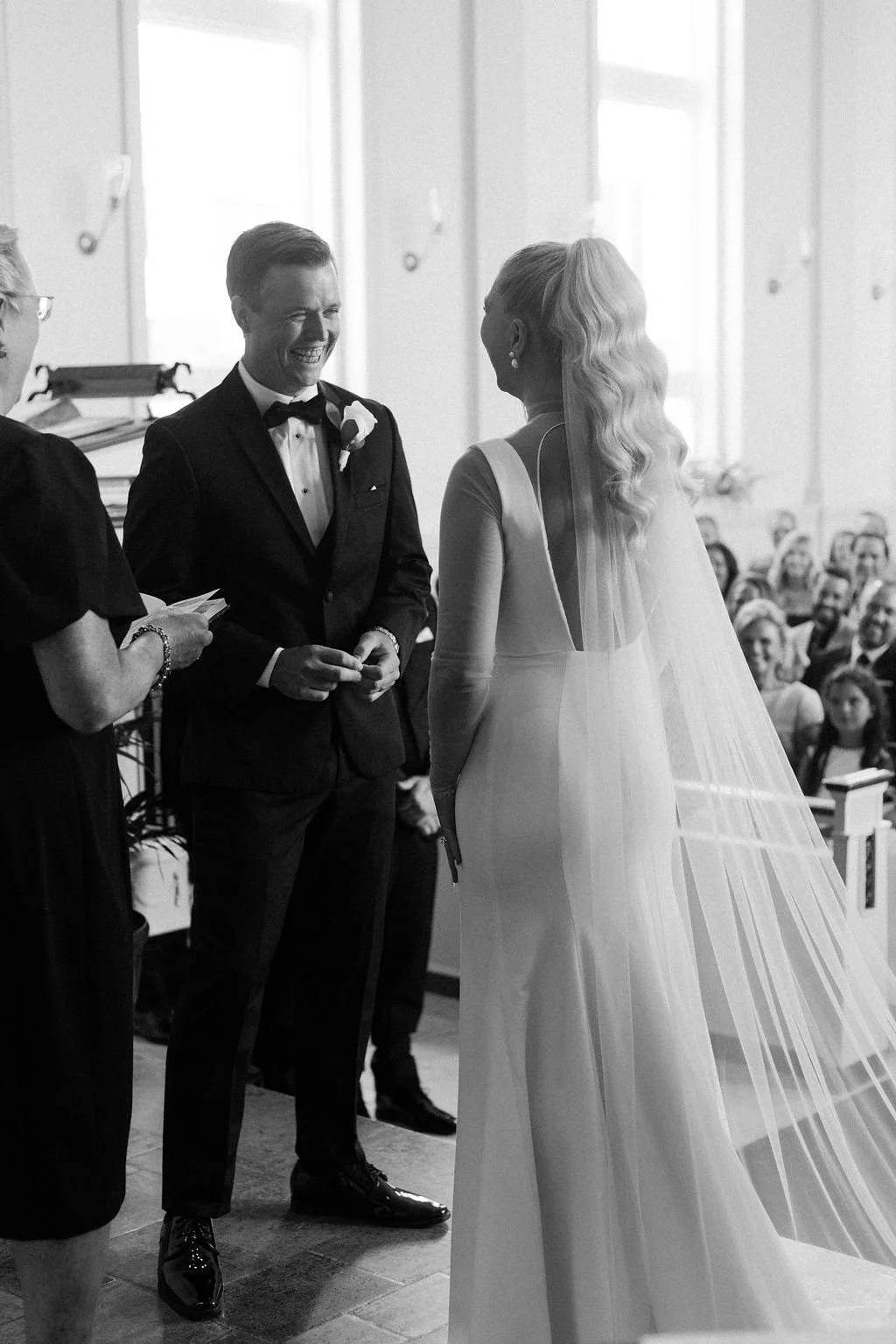 A black-and-white photo of a wedding ceremony showing a couple exchanging vows. The groom is smiling and dressed in a tuxedo, and the bride in a white wedding gown with a long veil. Guests are seated in the background.