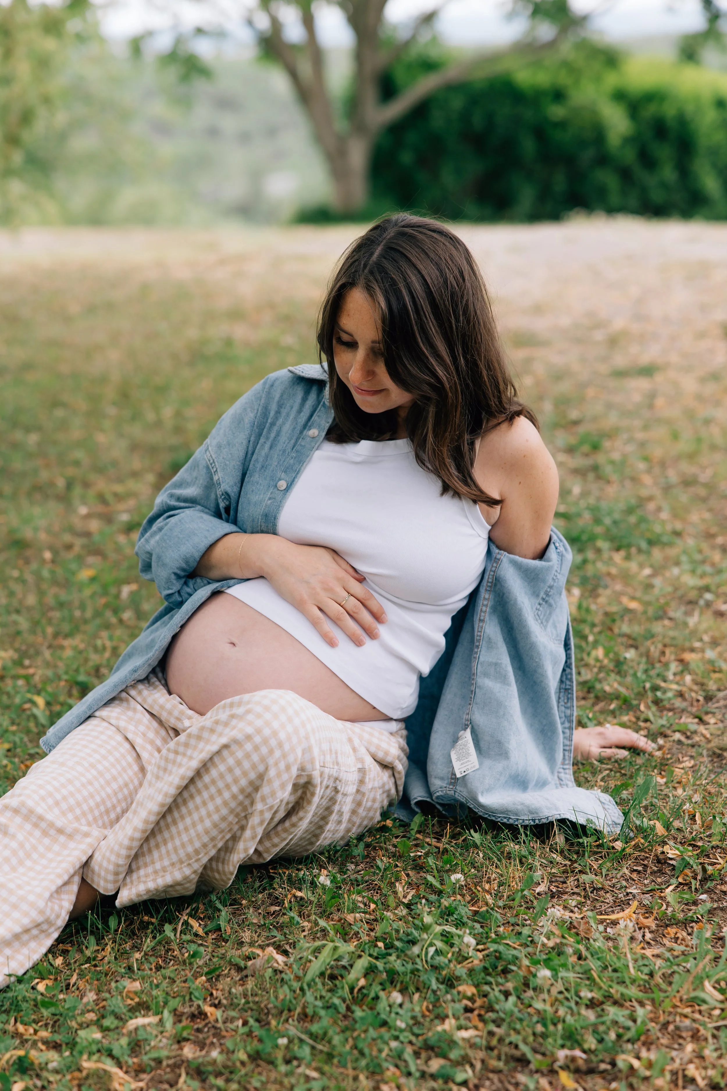 A woman with dark hair and light skin, wearing a denim jacket and white dress, sits outdoors on grass with a man with light skin, blonde hair, a beige checkered outfit, and plaid pants. The woman has her hand on her pregnant belly with her eyes close