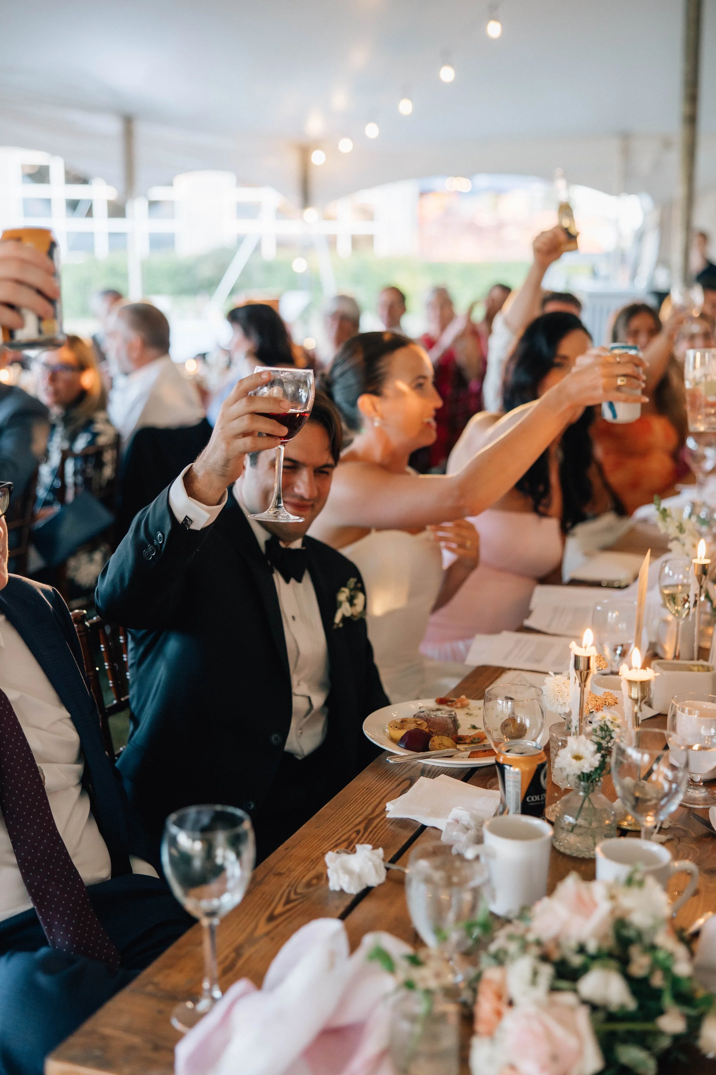 A large group of people celebrating at a wedding reception, seated at a long table with food, drinks, and floral decorations in a tented outdoor venue.