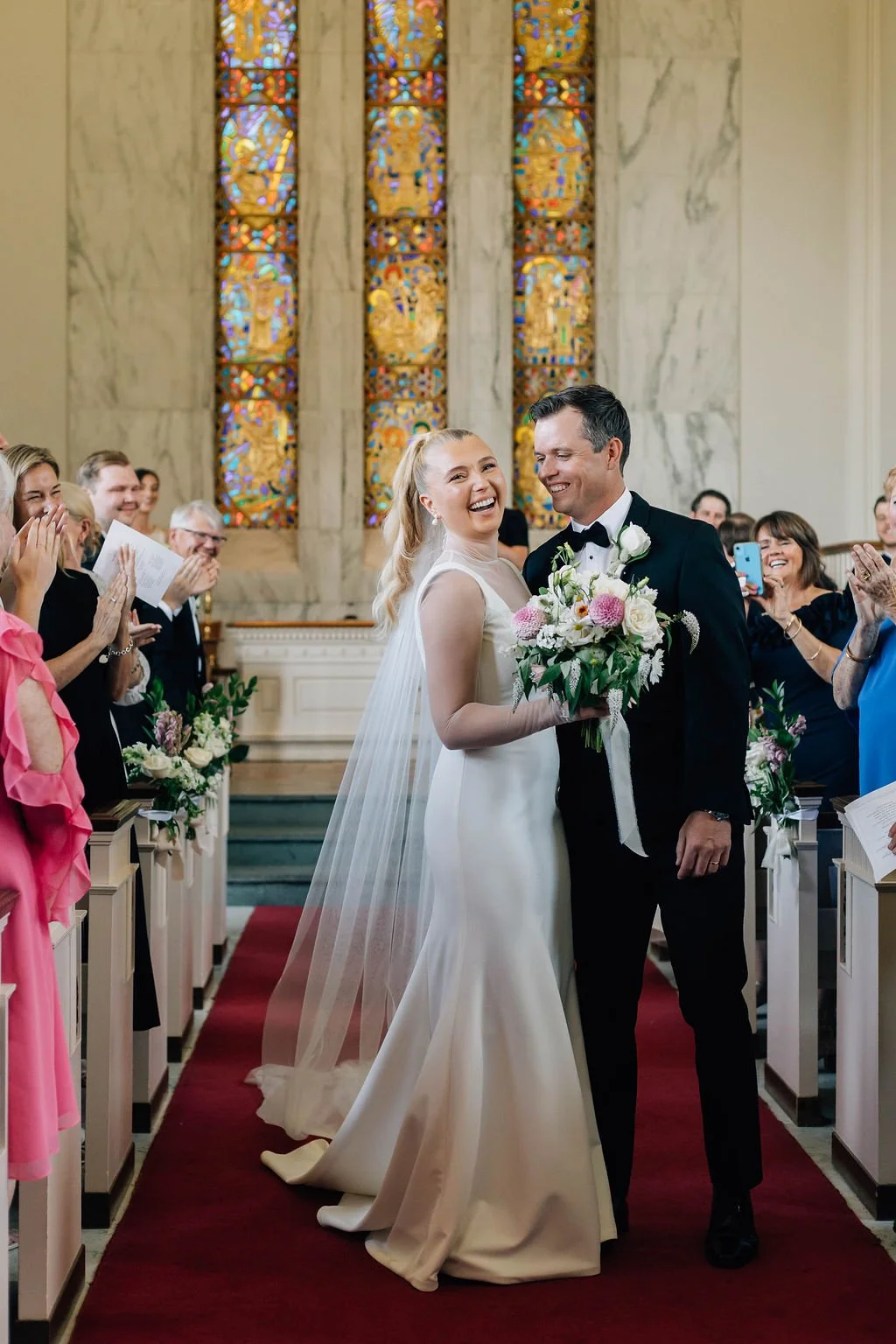 A newlywed couple stands at the altar in a church, smiling and holding a bouquet of flowers, surrounded by guests who are clapping and smiling.