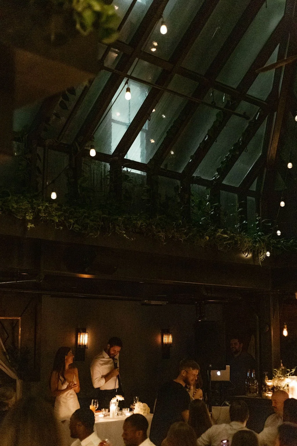 A dimly lit indoor event with a man and woman speaking at a table, surrounded by seated guests, and a glass ceiling with hanging lights.
