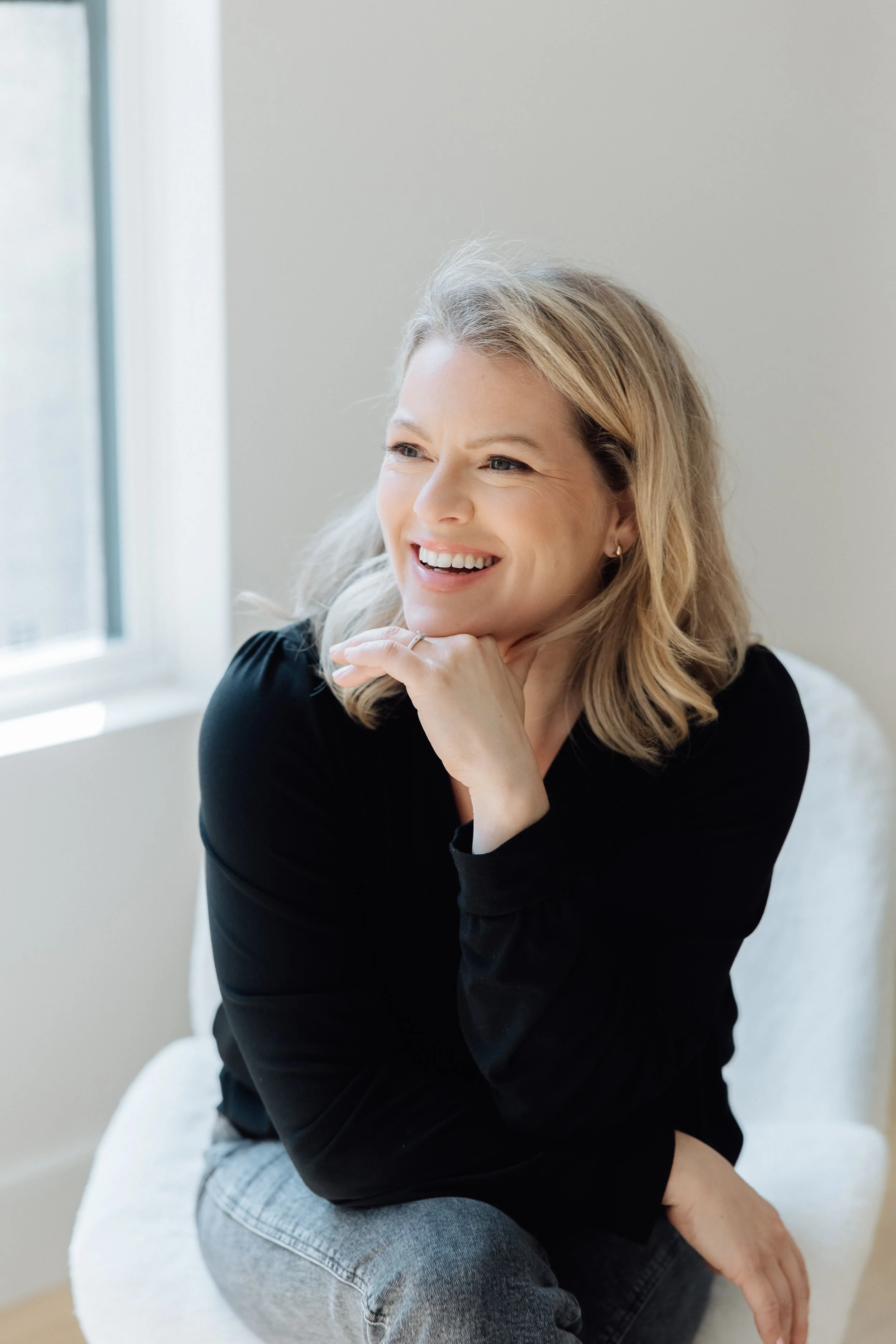 A woman with blonde hair smiling, sitting on a white chair near a window, wearing a black top and gray pants, with her right hand resting under her chin.
