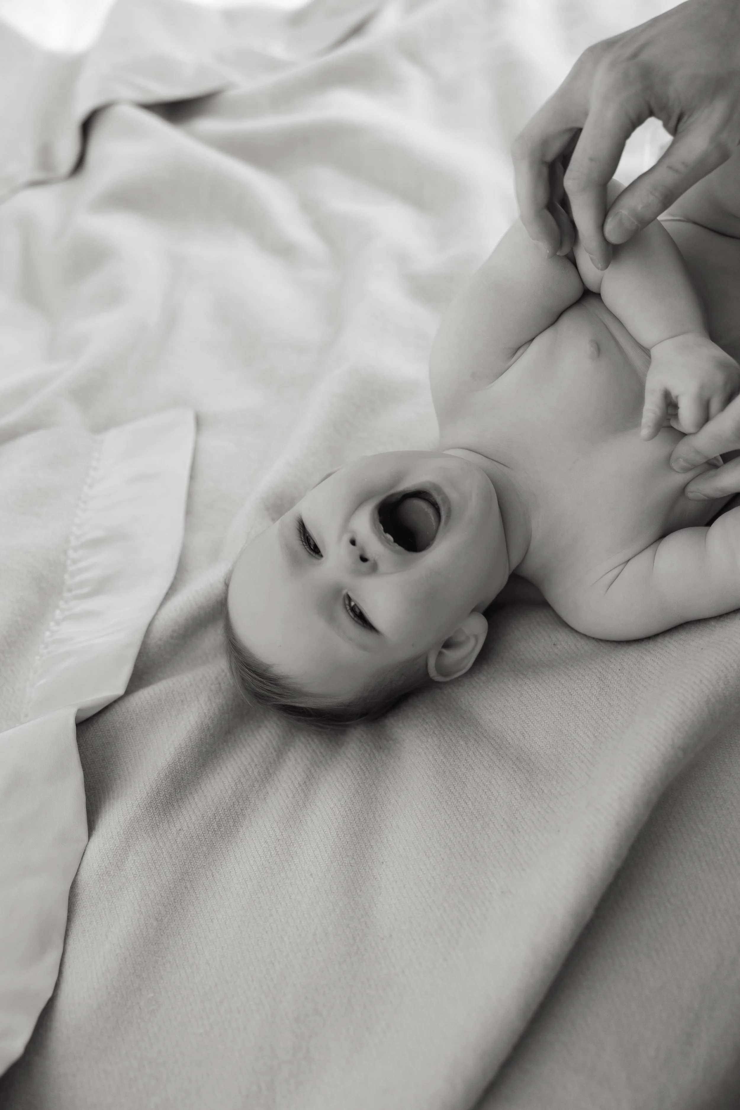 A baby lying on a bed with a blanket, yawning or crying, while an adult's hand gently holds the baby's foot.