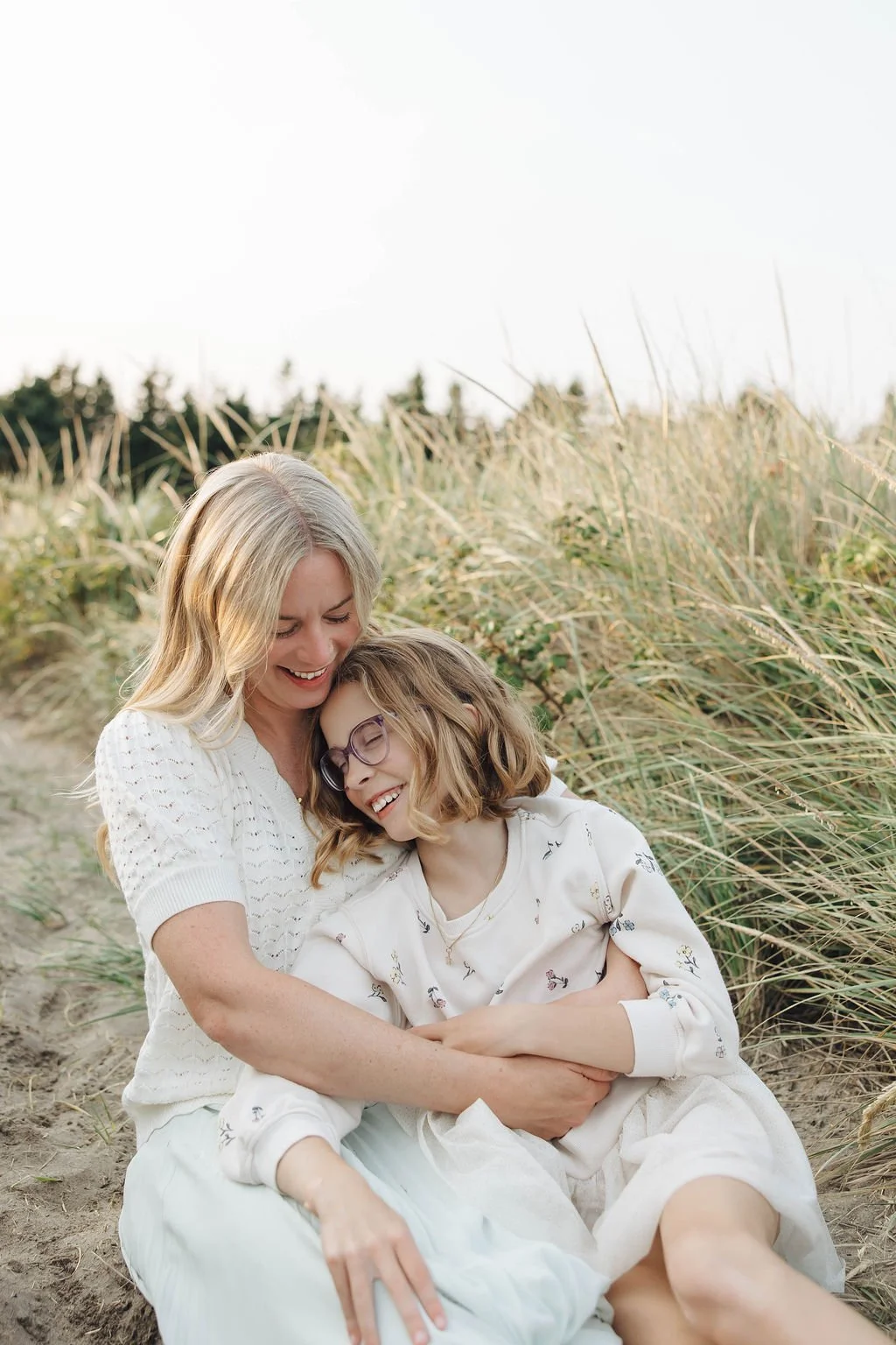 A joyful woman and girl sitting on sandy ground in a grassy field, hugging and smiling.