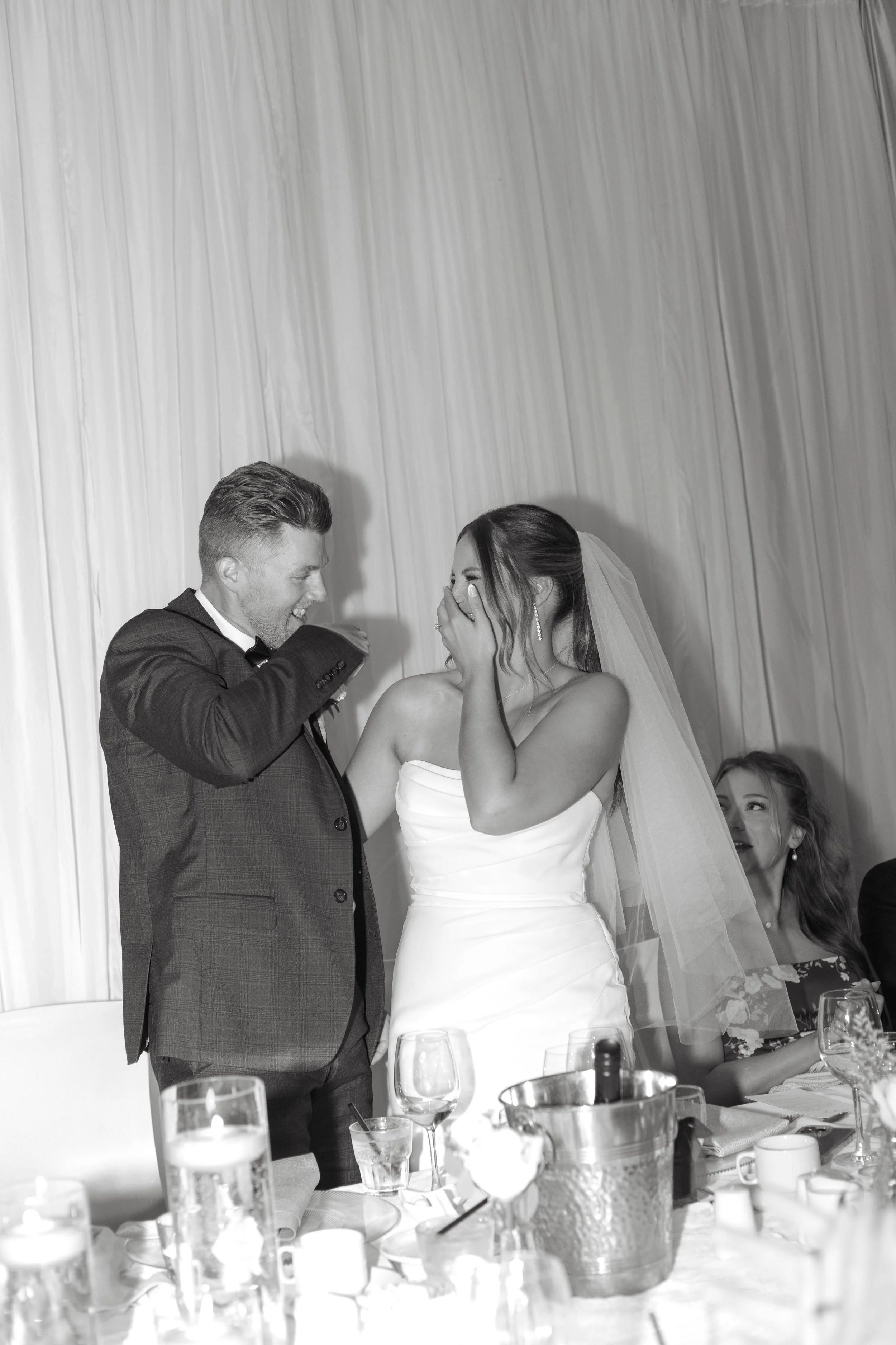 A wedding reception scene in black and white, with a bride and groom standing close together. The bride is covering her mouth with her hand, smiling, and dressed in a strapless wedding gown with a veil. The groom is wearing a suit with a bow tie and 