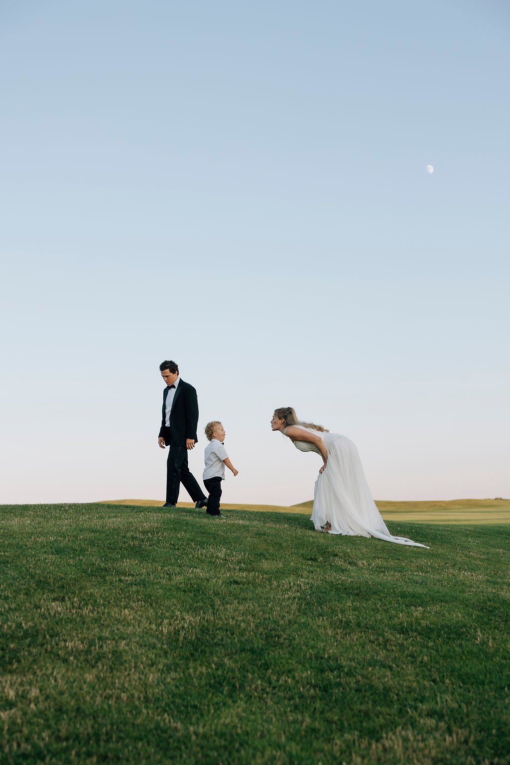 A man in a tuxedo, a woman in a wedding dress, and a young boy are outdoors on a grassy hill under a clear sky with the moon visible. The woman is bending forward and facing the boy, while the man is standing and looking down at them.