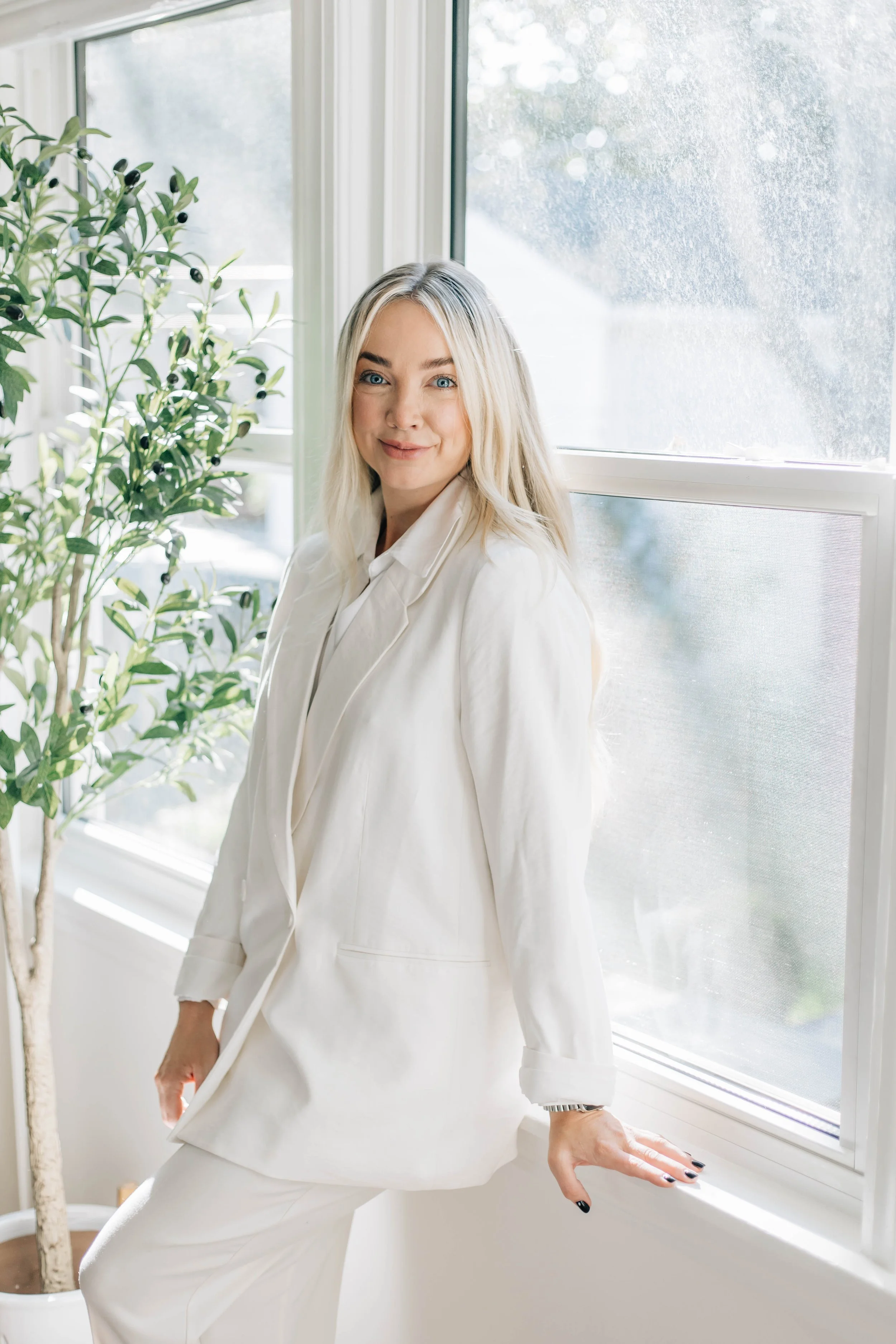 A woman wearing a white blazer and pants standing near a window with greenery outside. She has blonde hair and is smiling softly.