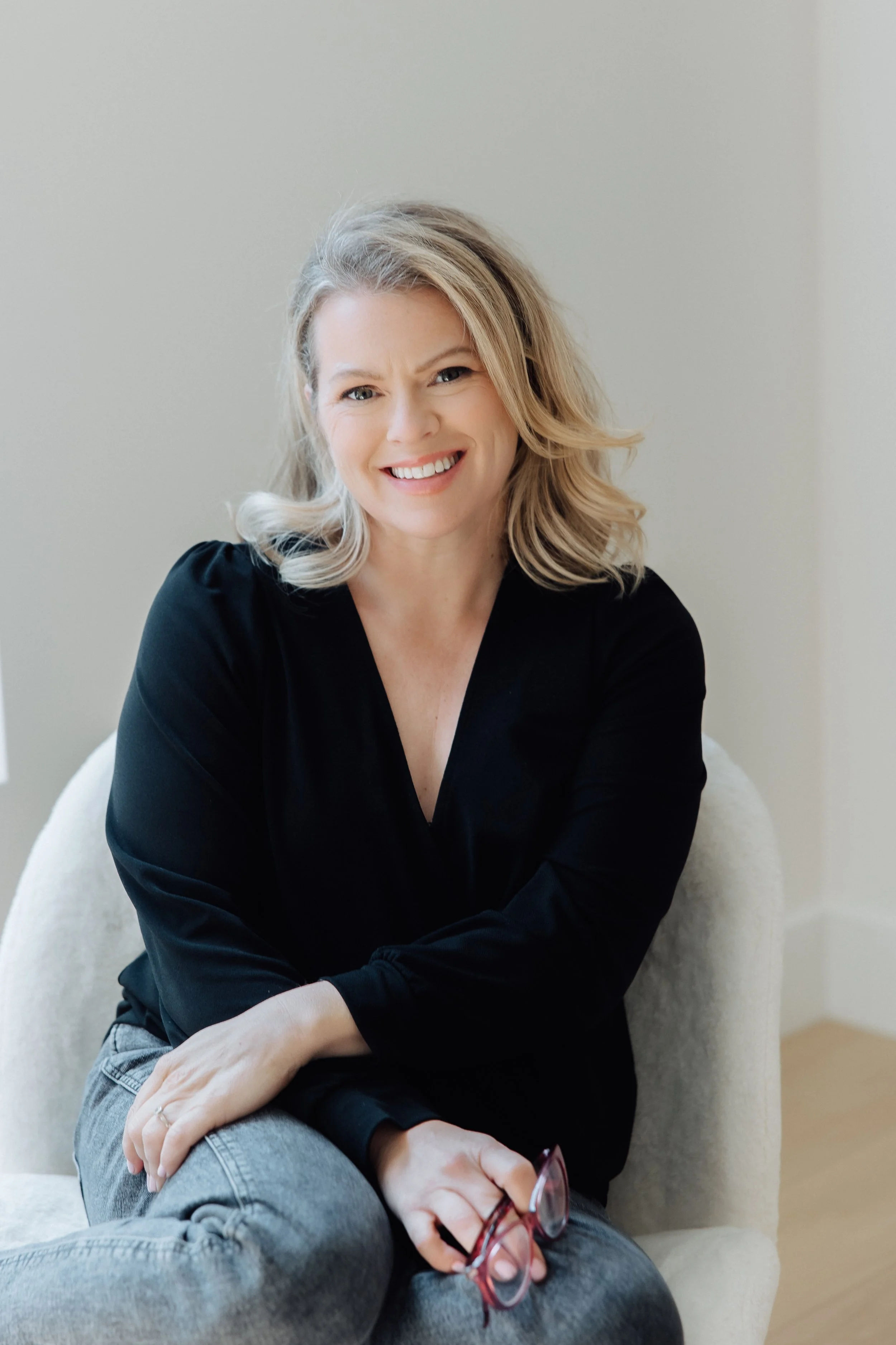 A smiling woman with blonde, wavy hair sitting on a beige chair, wearing a black top and gray jeans, holding glasses in her right hand, against a plain light-colored wall.