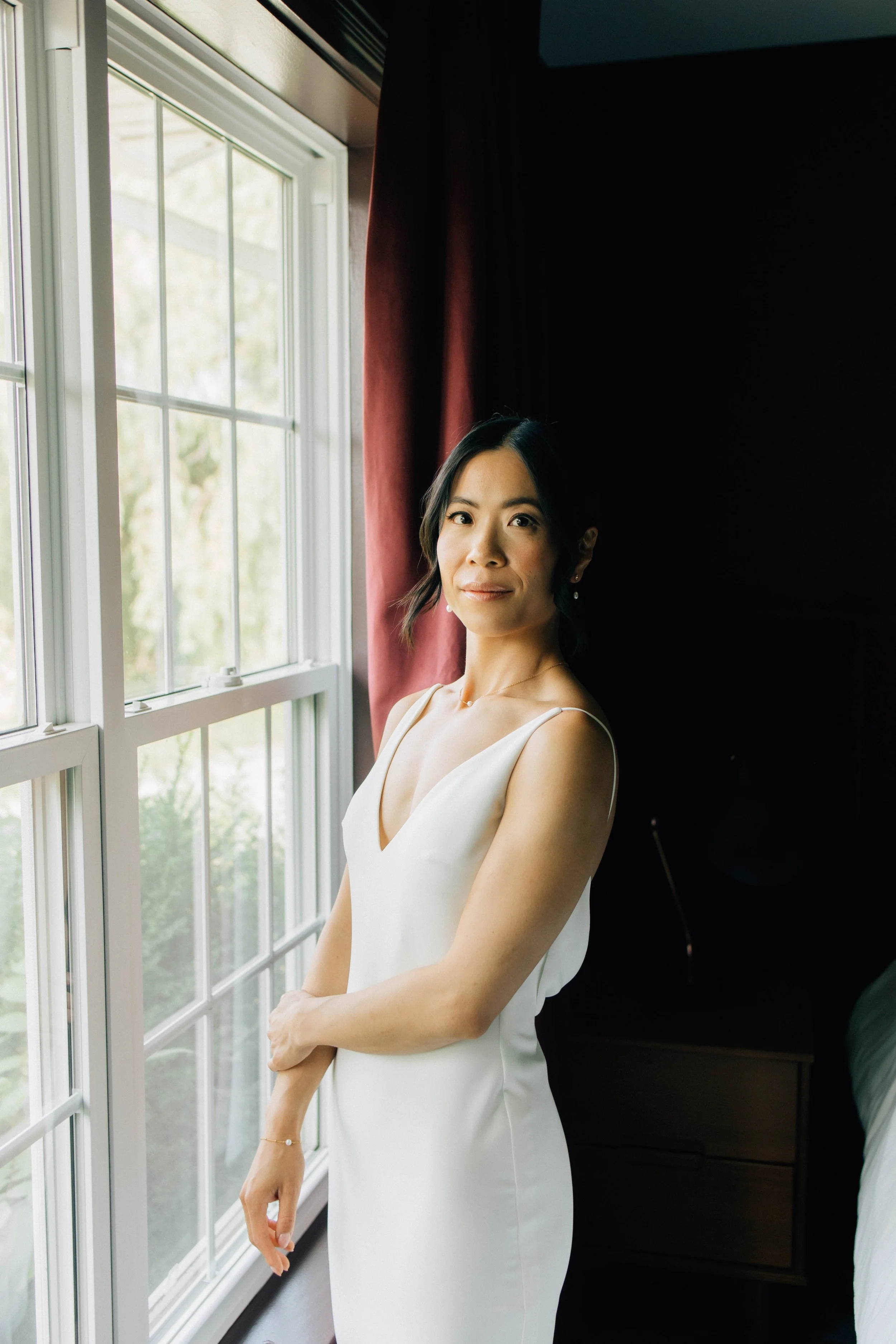 A woman in a white satin dress standing near a window with sunlight, dark curtains, and a black piece of furniture behind her.