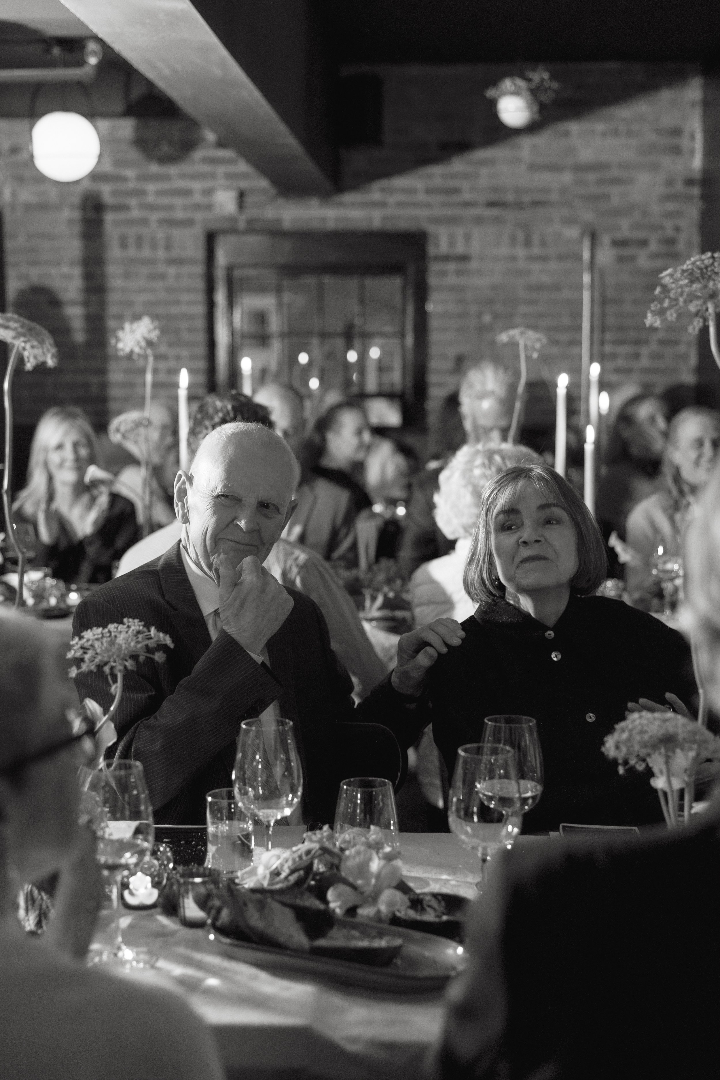 Black and white photo of a formal dinner event with people seated at a table, including an elderly man in a suit and a woman. The table has glasses, flowers, and candles, and the background features a brick wall and window.