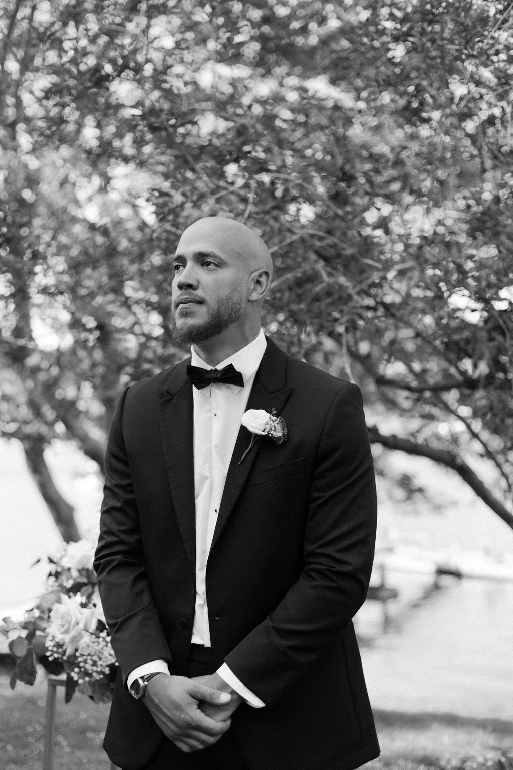 A man in a tuxedo with a bow tie and boutonnière stands outdoors, looking to the left, with trees in the background.