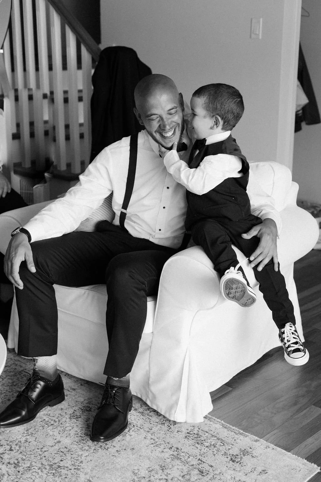 A man and young boy dressed in formal attire sharing a joyful moment indoors. The boy is playfully touching the man's face while sitting on a white armchair.