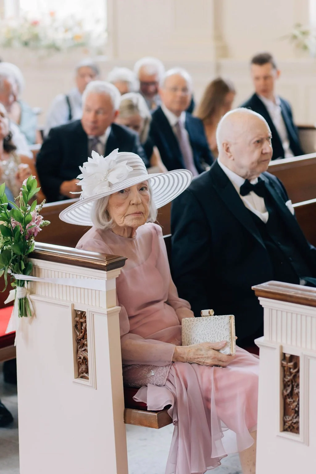 Older woman in a pink dress and wide-brimmed hat sitting in a church pew during a wedding ceremony, holding a white clutch purse, with other guests seated behind her.