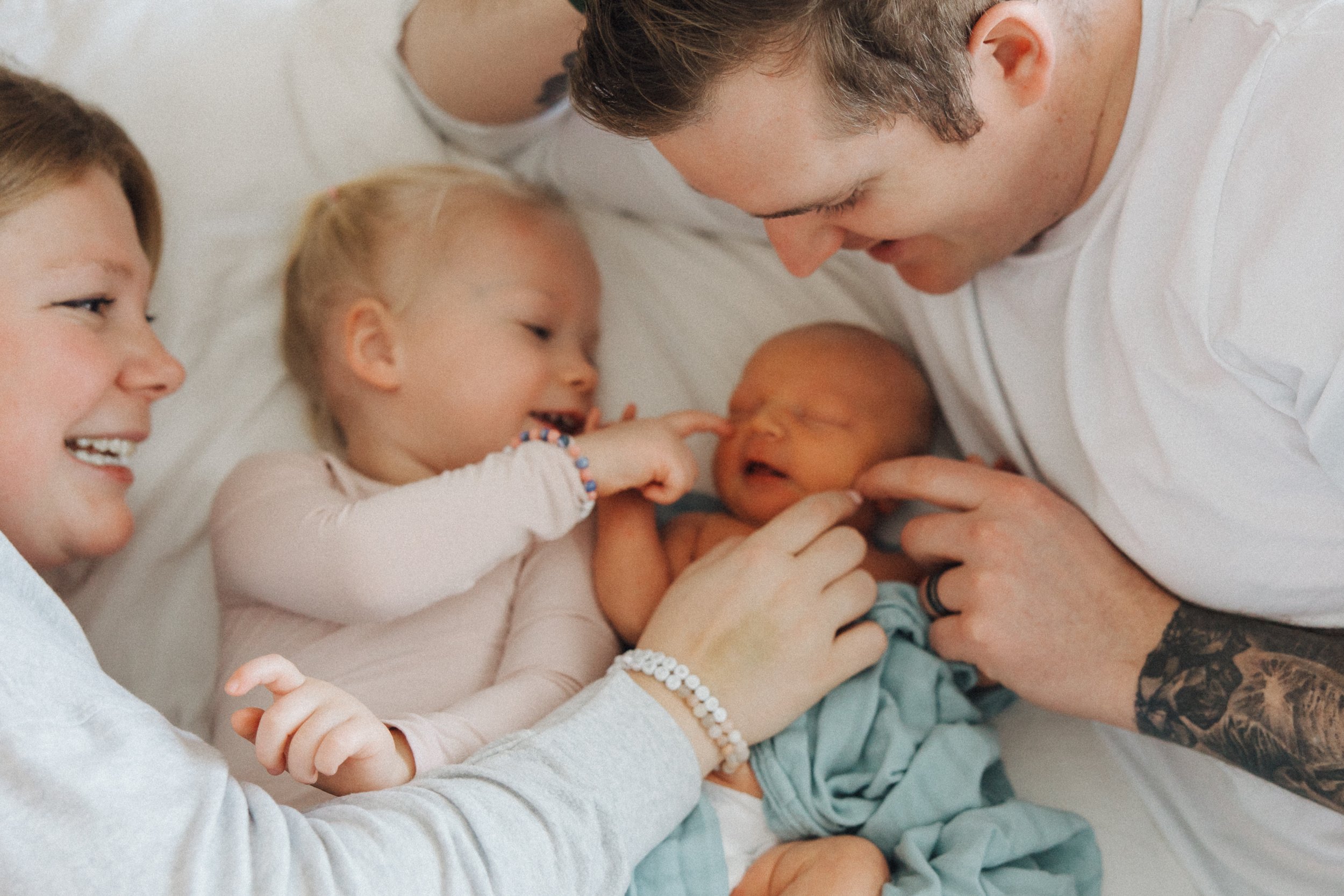 A family of four laying on a bed, celebrating the birth of a newborn baby. The father is gently holding the baby, while the mother, and two young children are smiling and touching the newborn.