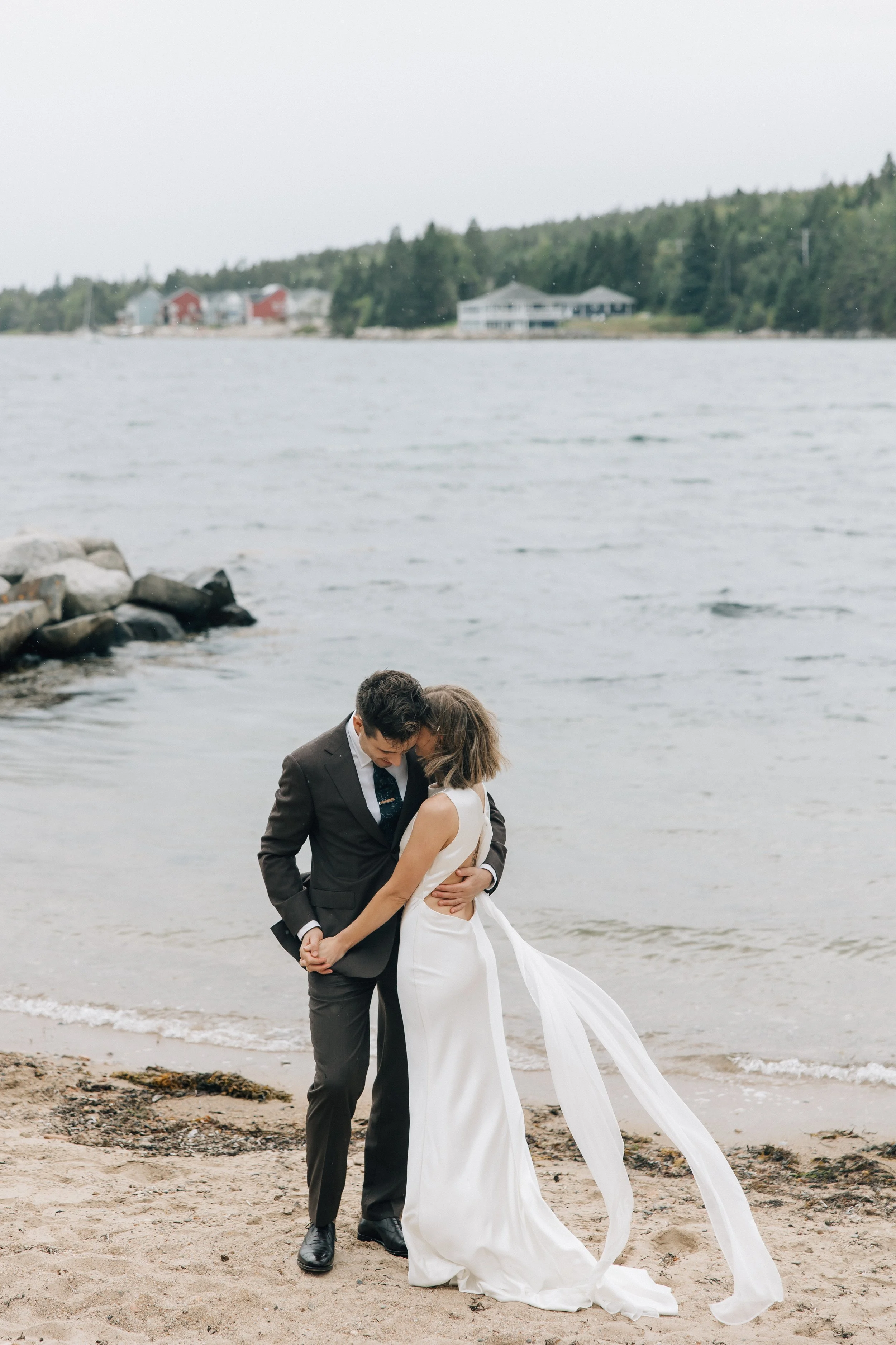 A bride and groom embrace on a beach with water and houses in the background, under an overcast sky.