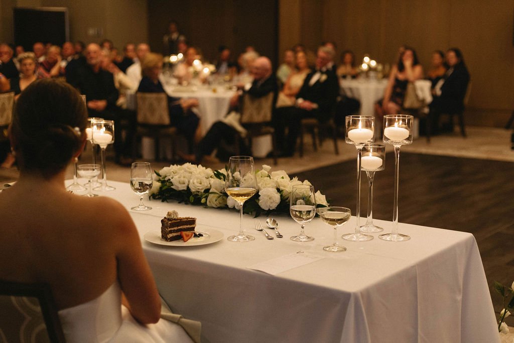 Bride sitting at a head table during a wedding reception with a wedding cake slice, wine, and floral centerpieces, while guests watch in the background.