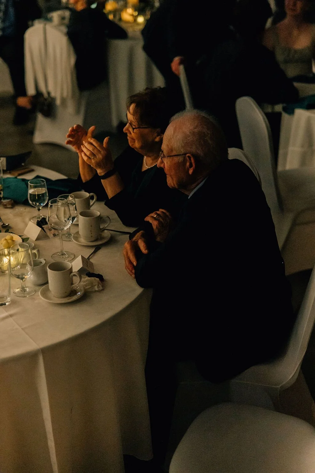 An elderly man and woman seated at a formal dinner table, clapping, surrounded by cups, glasses, and silverware, in a dimly lit banquet hall.