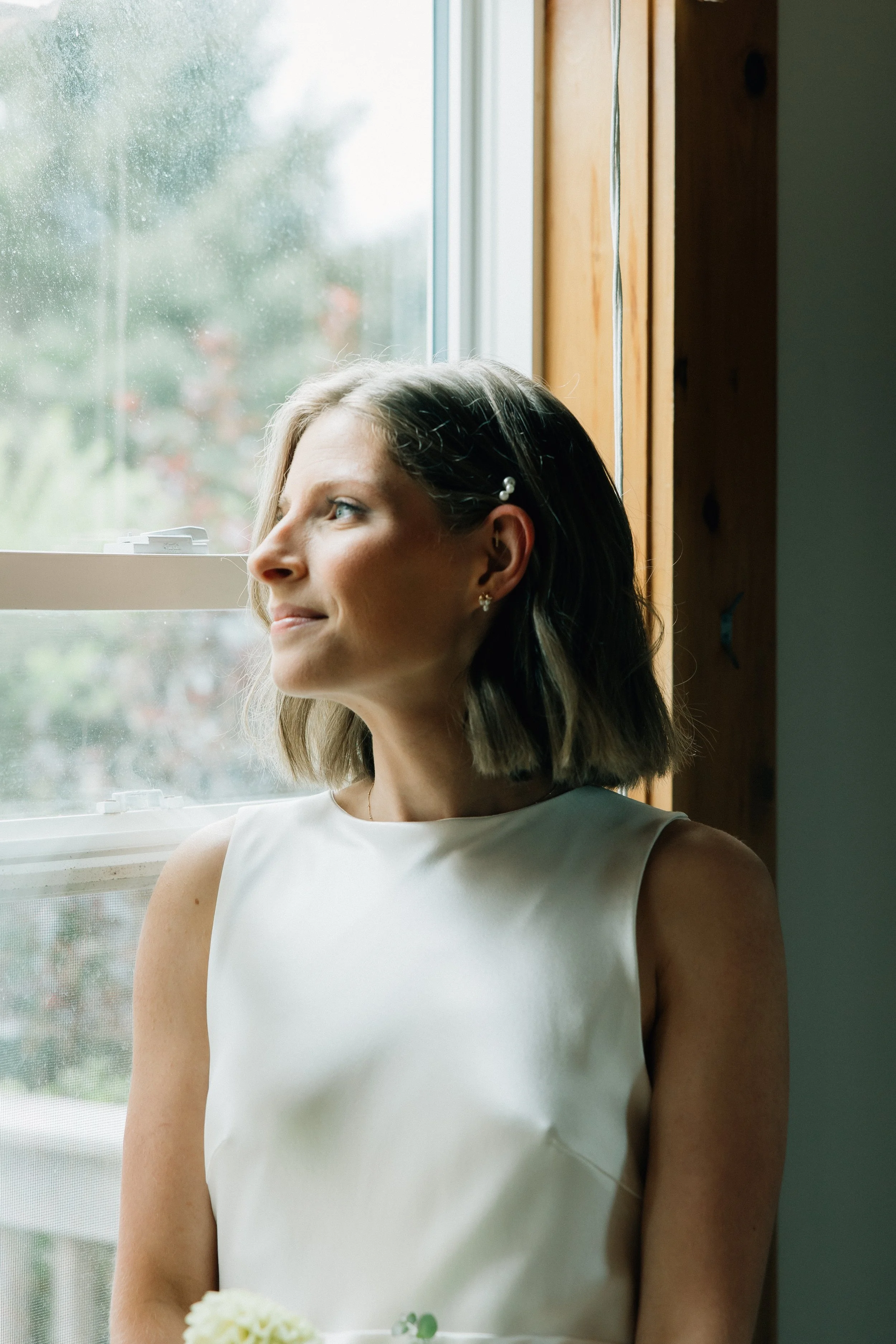 A woman with light skin and blonde hair, wearing a white sleeveless dress and pearl earrings, standing by a window with raindrops on the glass. She is looking out the window with a slight smile and appears to be holding a white flower in her hand.