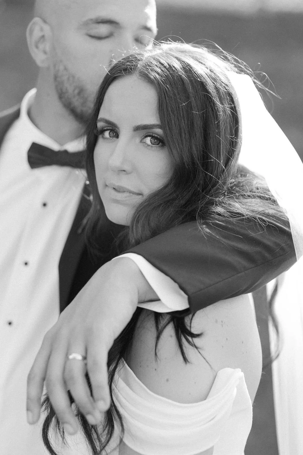 A black and white photograph of a bride with long dark hair, wearing an off-the-shoulder dress and a veil, posing with a groom who has a beard, dressed in a tuxedo with a bowtie; the groom is kissing the bride's temple.