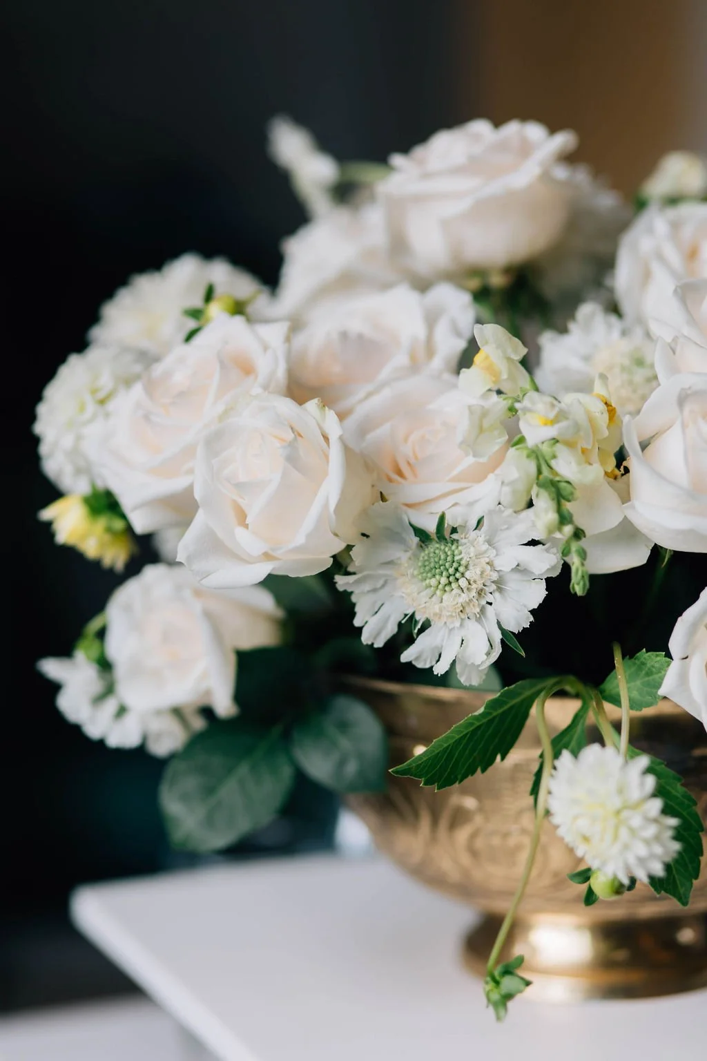 A gold-colored vase holding a bouquet of white roses, white daisies, and other white flowers with green leaves.