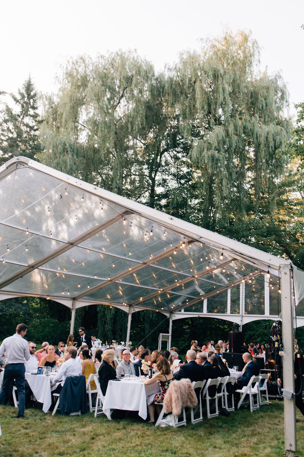 Outdoor wedding reception under a clear tent with string lights, surrounded by green trees and grass, guests seated at long tables.