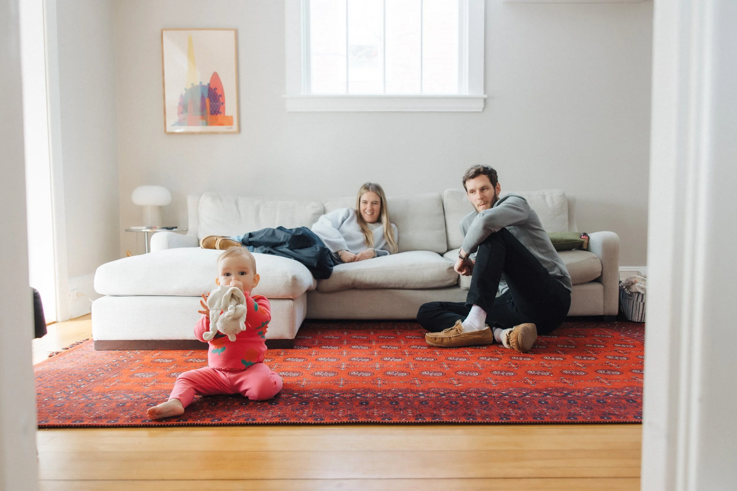 A family of three in a living room. A baby in pink pajamas sits on a red patterned rug in the foreground holding a stuffed animal. Behind, a woman lies on a white sofa smiling, while a man sits on the floor near the sofa looking towards the baby. The