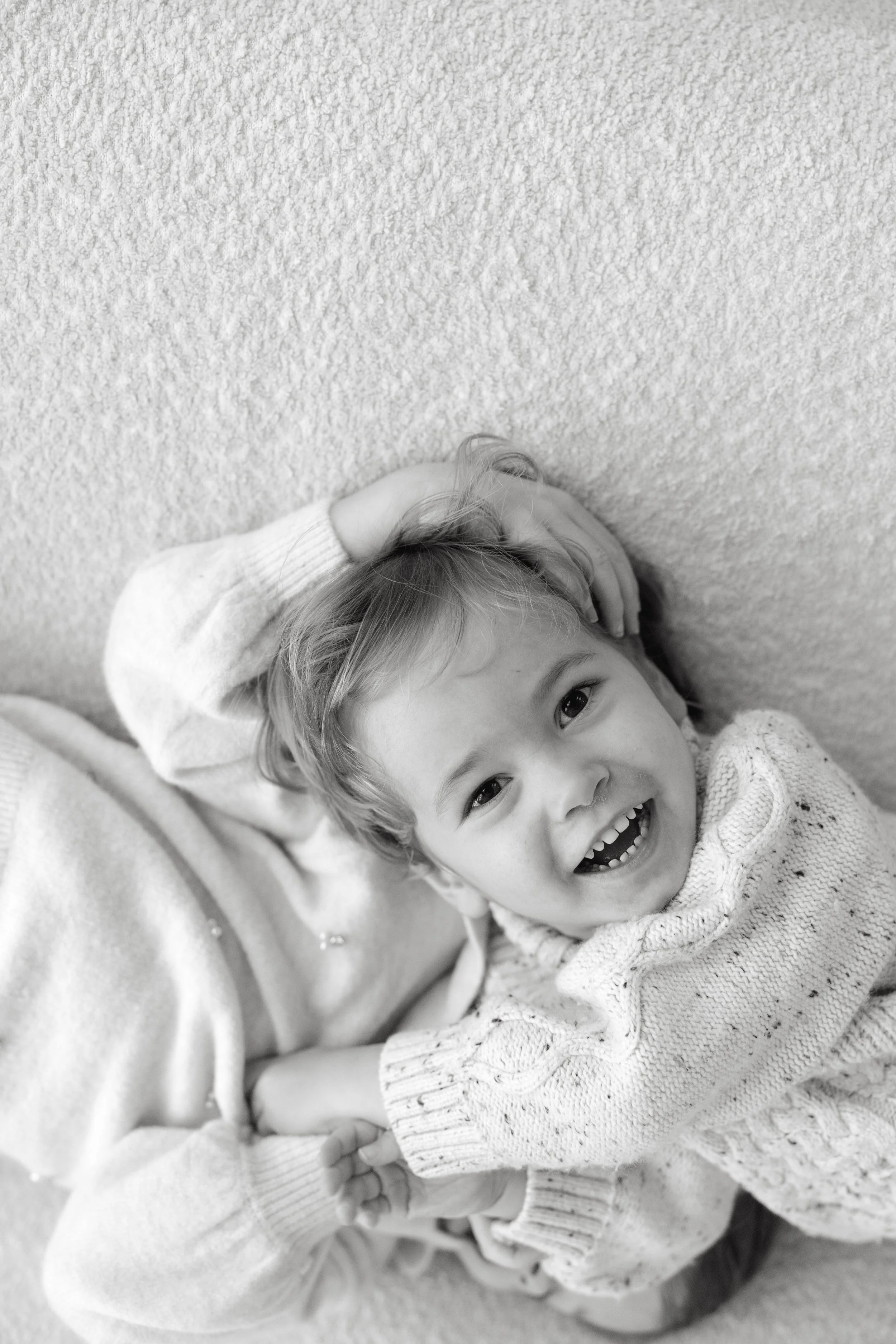 A young child with light hair lying on a textured surface, smiling widely with one hand on her head. She is wearing a patterned sweater.