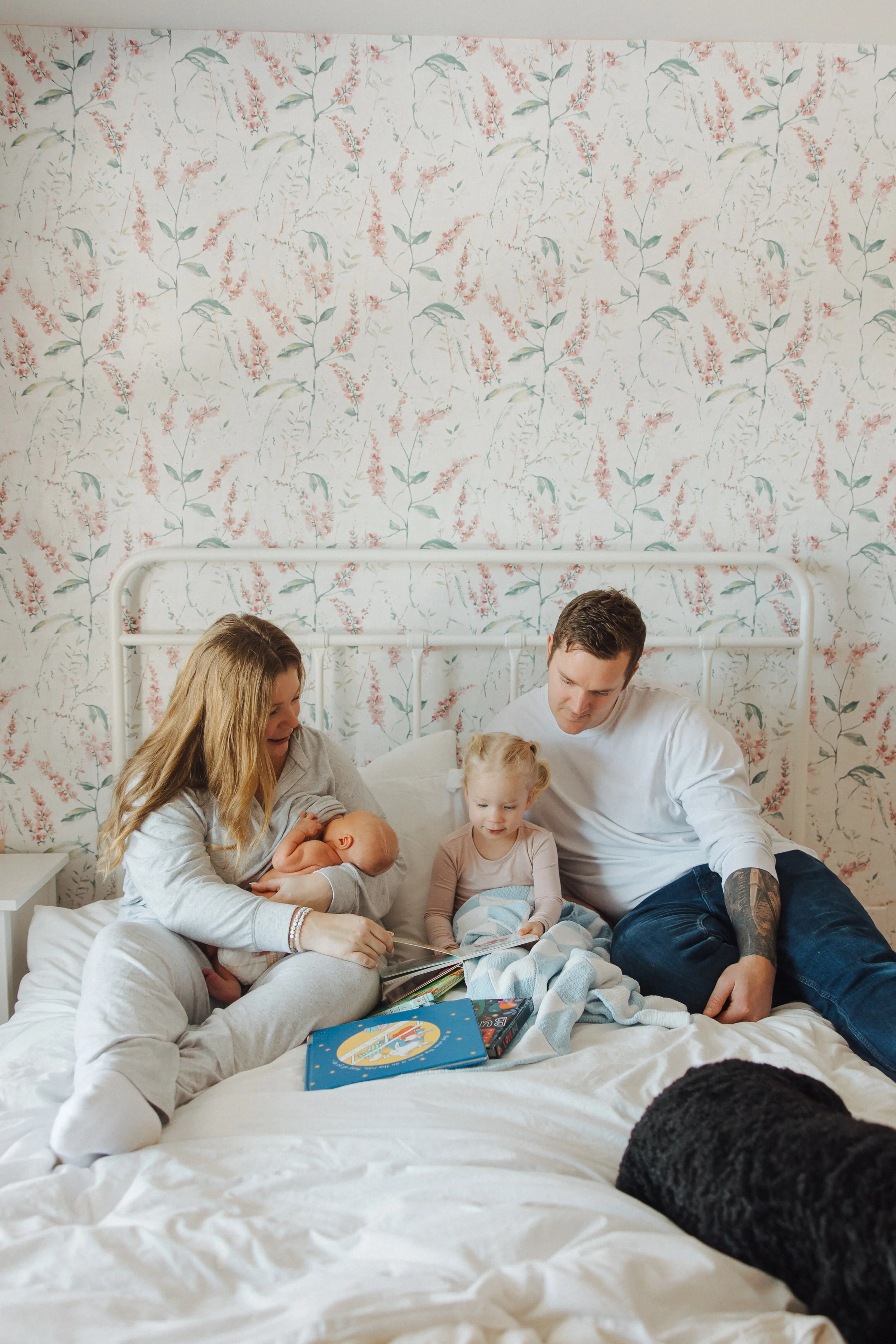 Family of four sitting on a bed with a floral wallpaper background, children and parents enjoying reading a book together.
