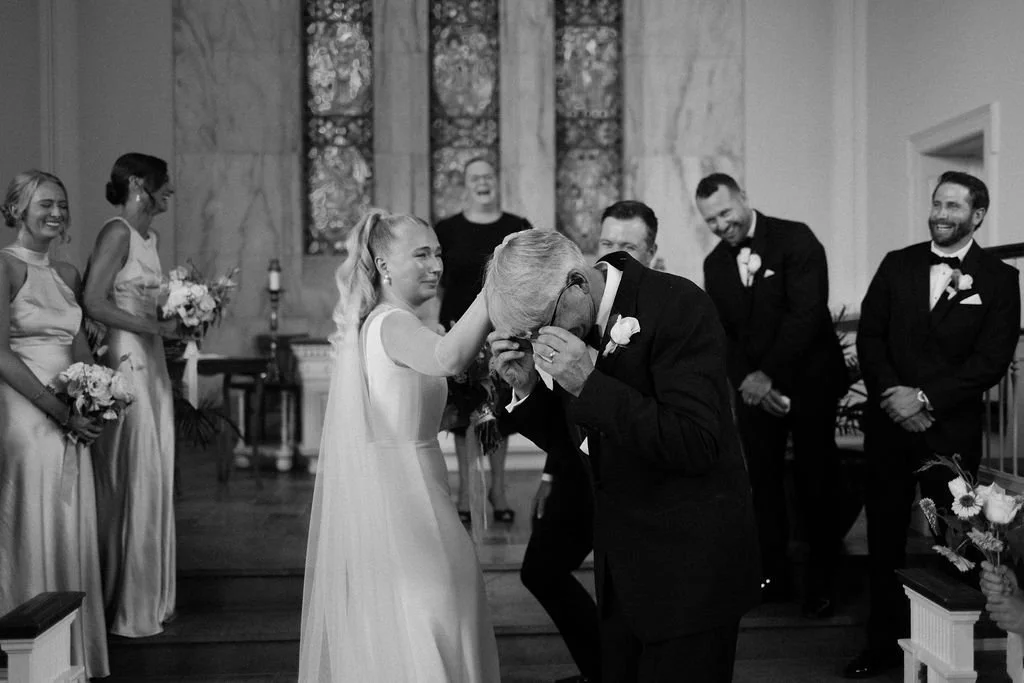 A bride and an elderly man are emotional, with the man wiping tears during a wedding ceremony in a church, surrounded by bridesmaids and groomsmen.