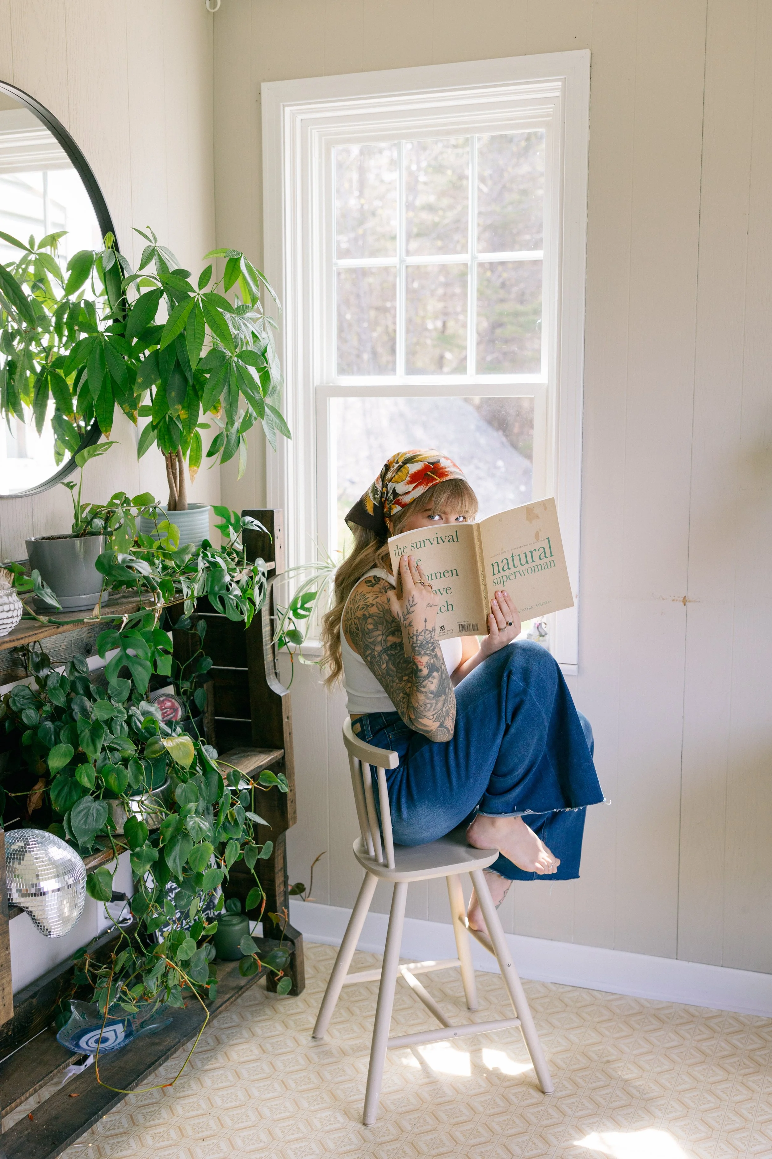 A woman with tattoos, wearing a white tank top, and blue jeans, sitting barefoot on a wooden chair in a sunlit room, holding a book titled "Natural Superwoman" in front of her face. The room has a large window with white trim, beige walls, and is dec