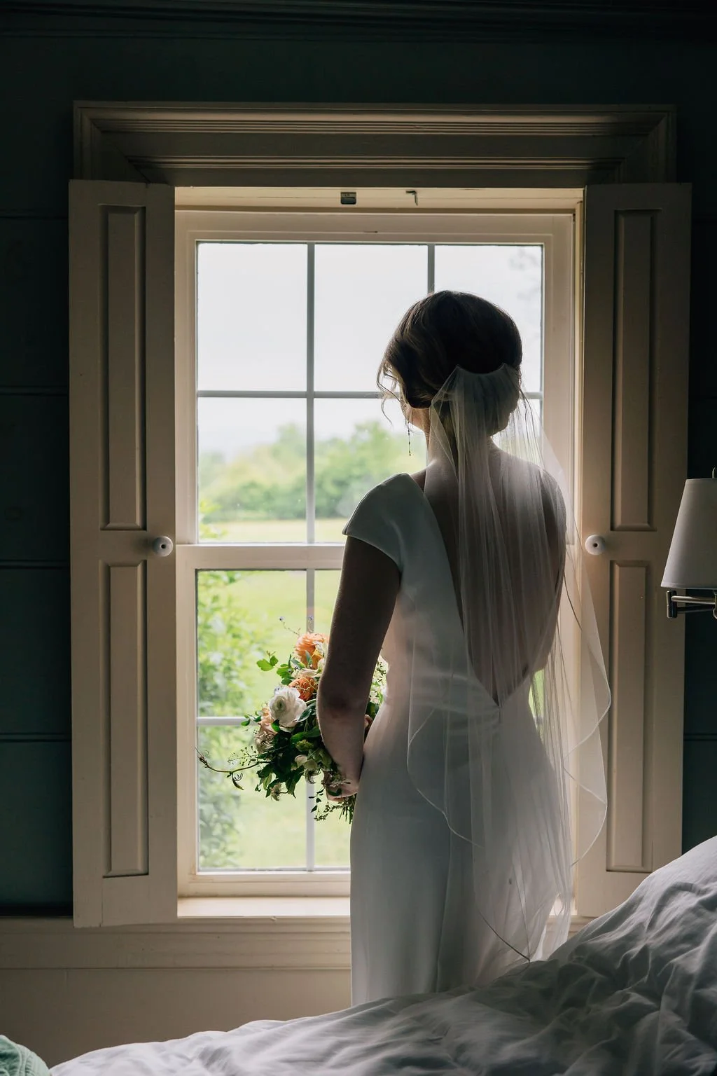A bride stands by a window holding a bouquet of flowers, looking outside, with a veil on her head and back to the camera.
