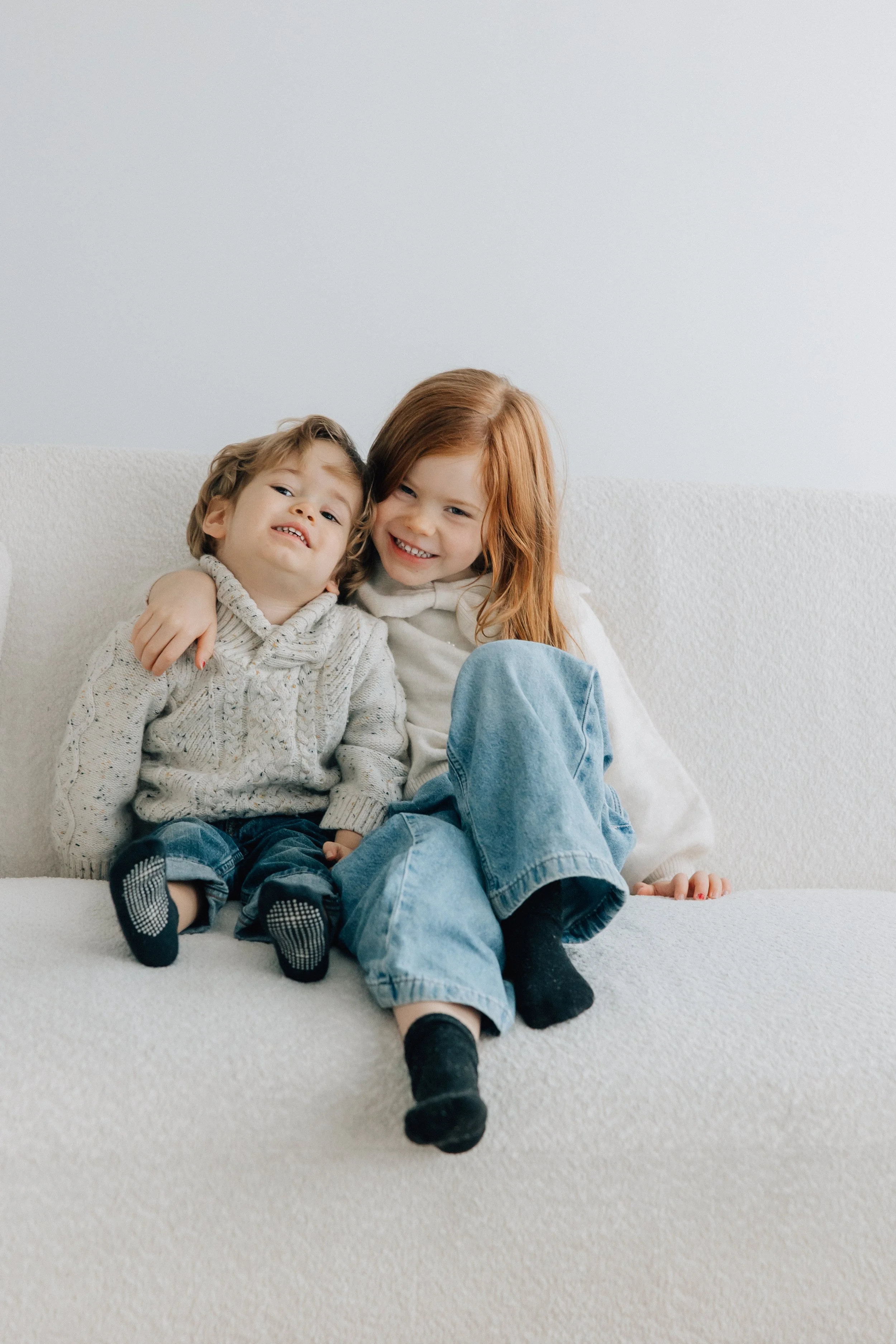 Two young children, a boy and a girl, sitting close together on a white couch, smiling and hugging each other in a cozy and friendly setting.