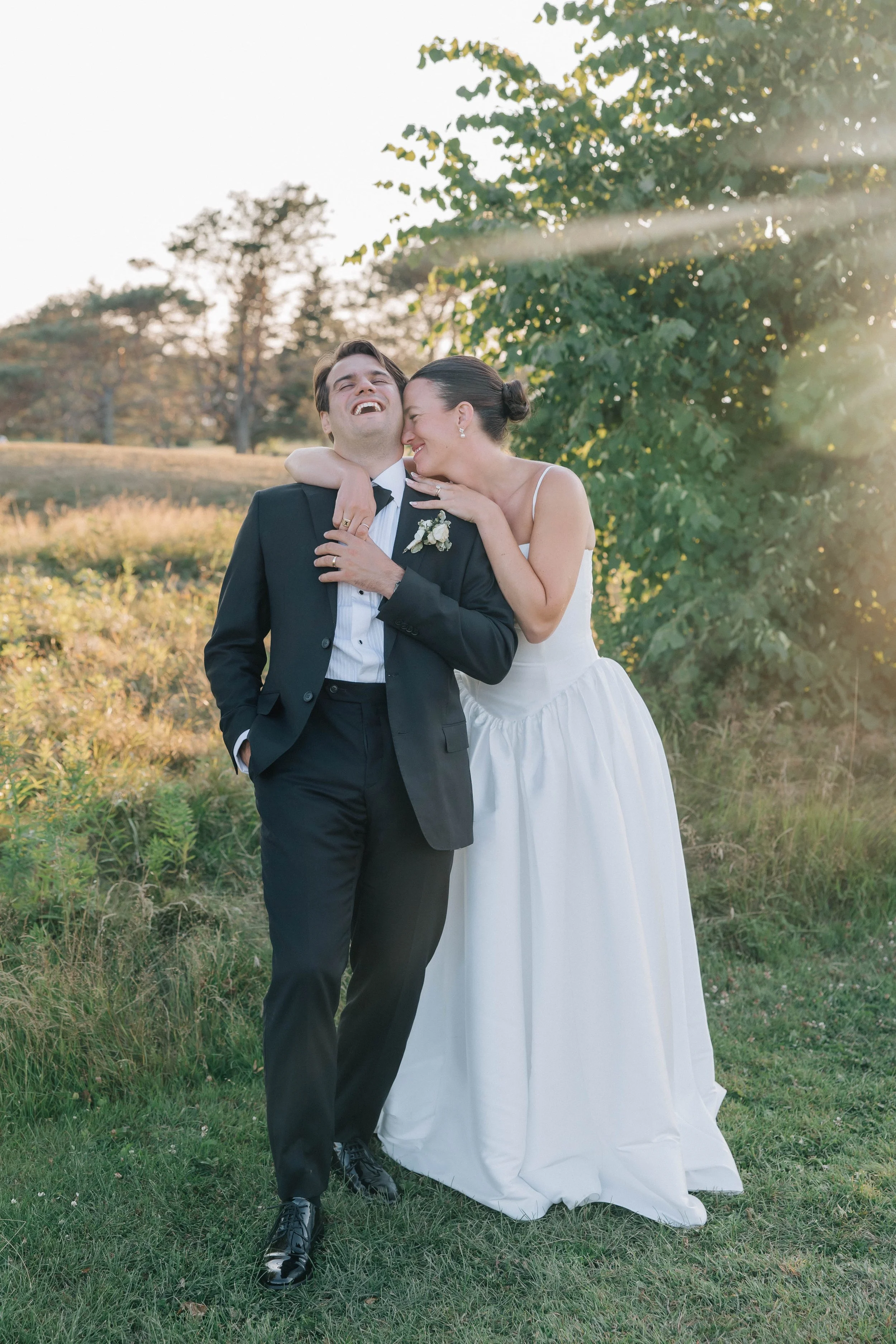 A newlywed couple standing in a field with trees in the background, sharing a joyful moment as they smile and laugh together on their wedding day.