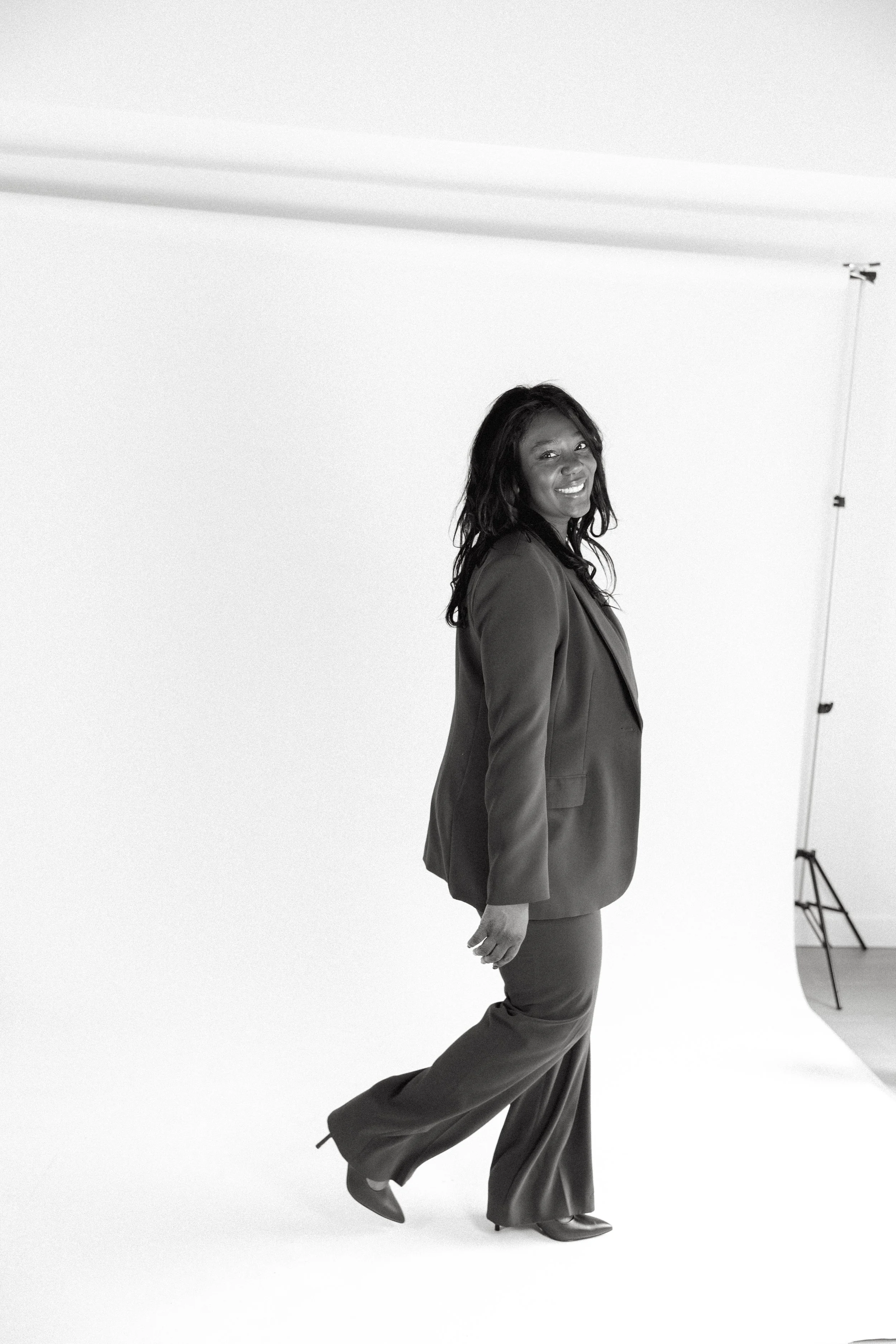 A smiling woman in a formal suit posing in a photography studio with a white backdrop and studio equipment.