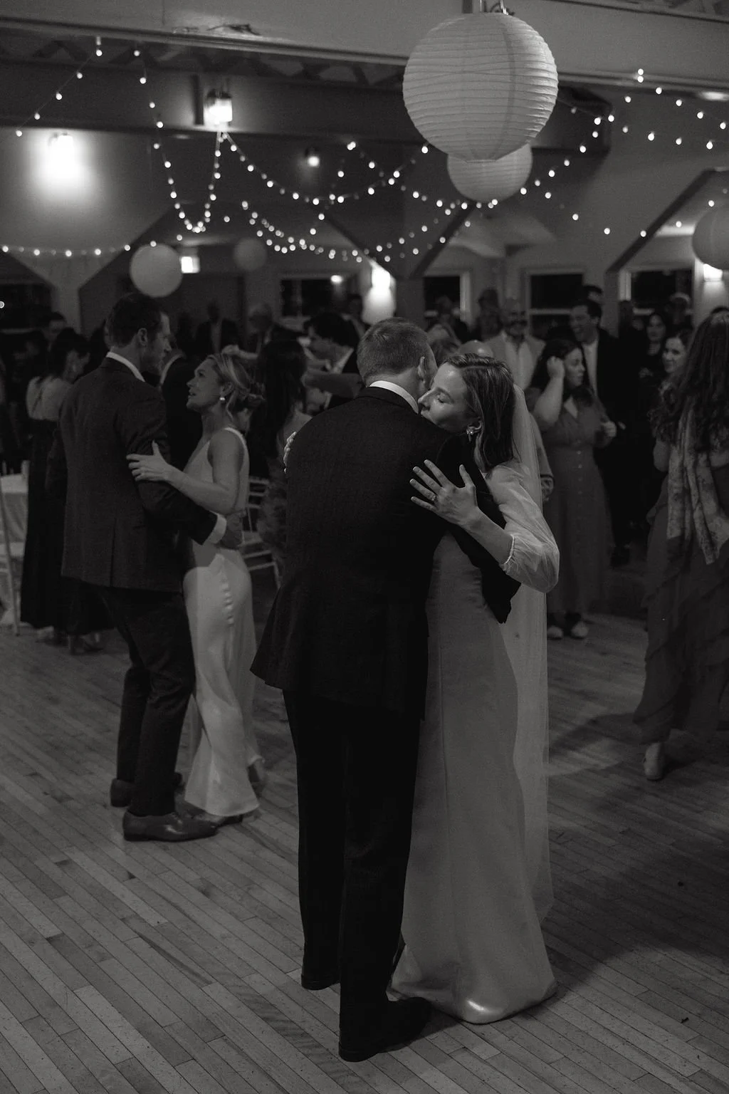 A black and white photo of a wedding reception with couples dancing, including a bride and groom sharing a dance in the foreground, decorated with hanging paper lanterns and string lights.