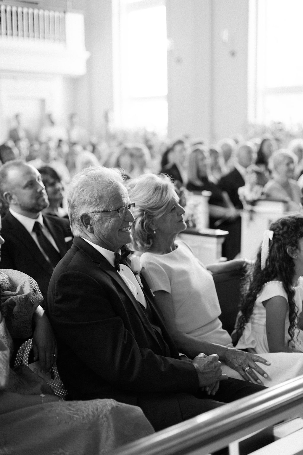 An elderly man with glasses in a tuxedo smiling and holding hands with an elderly woman in a white dress, sitting in a church or formal event, surrounded by a crowd of seated people.