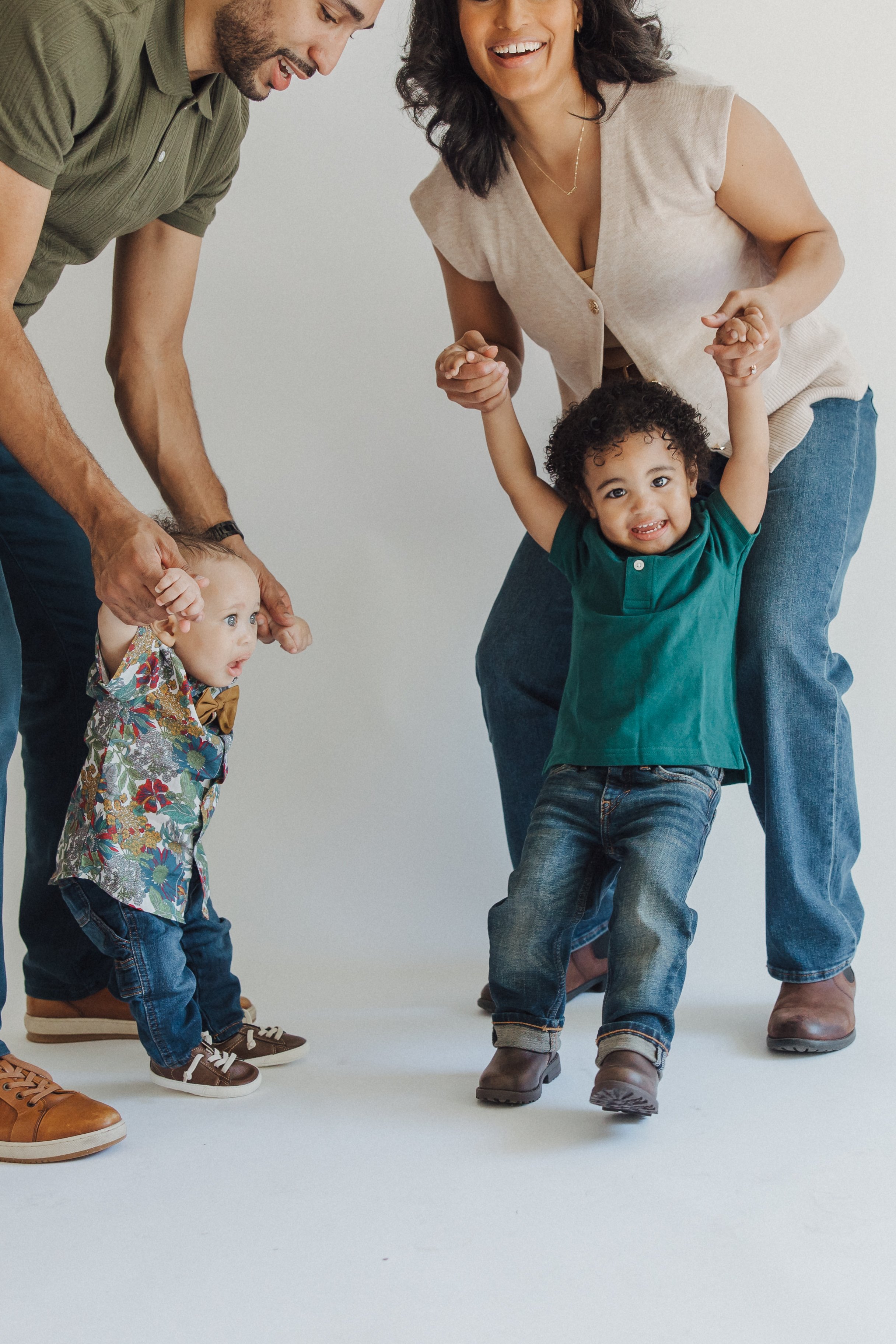 A diverse family of four, including two adults and two young children, enjoying a moment together. The adults are holding the children's hands, lifting them slightly, smiling and laughing. The children appear happy and engaged, with one child dressed