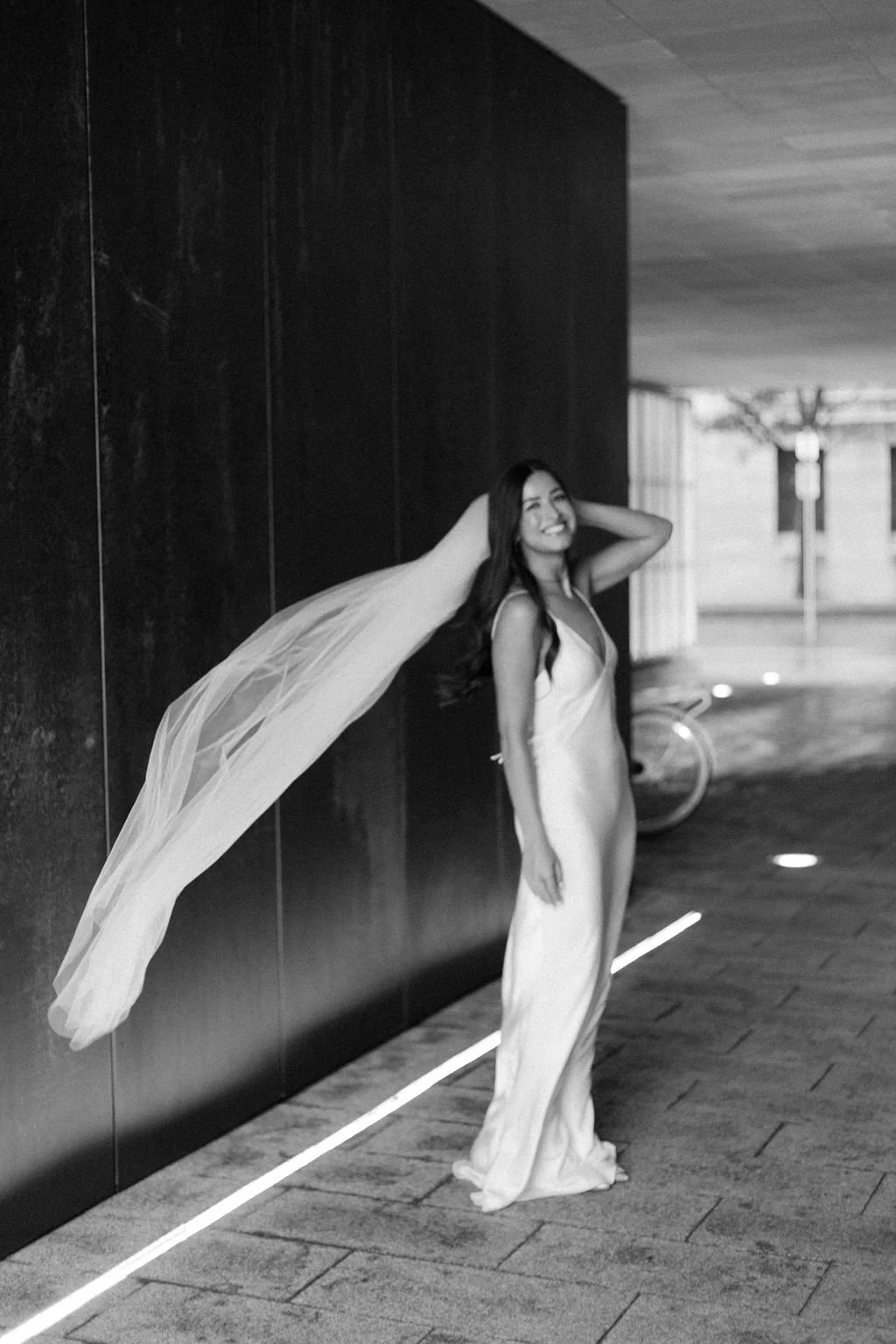 A woman in a wedding dress, smiling and posing outdoors at night, holding her veil with one hand behind her head, standing against a dark wall with a bicycle in the background.