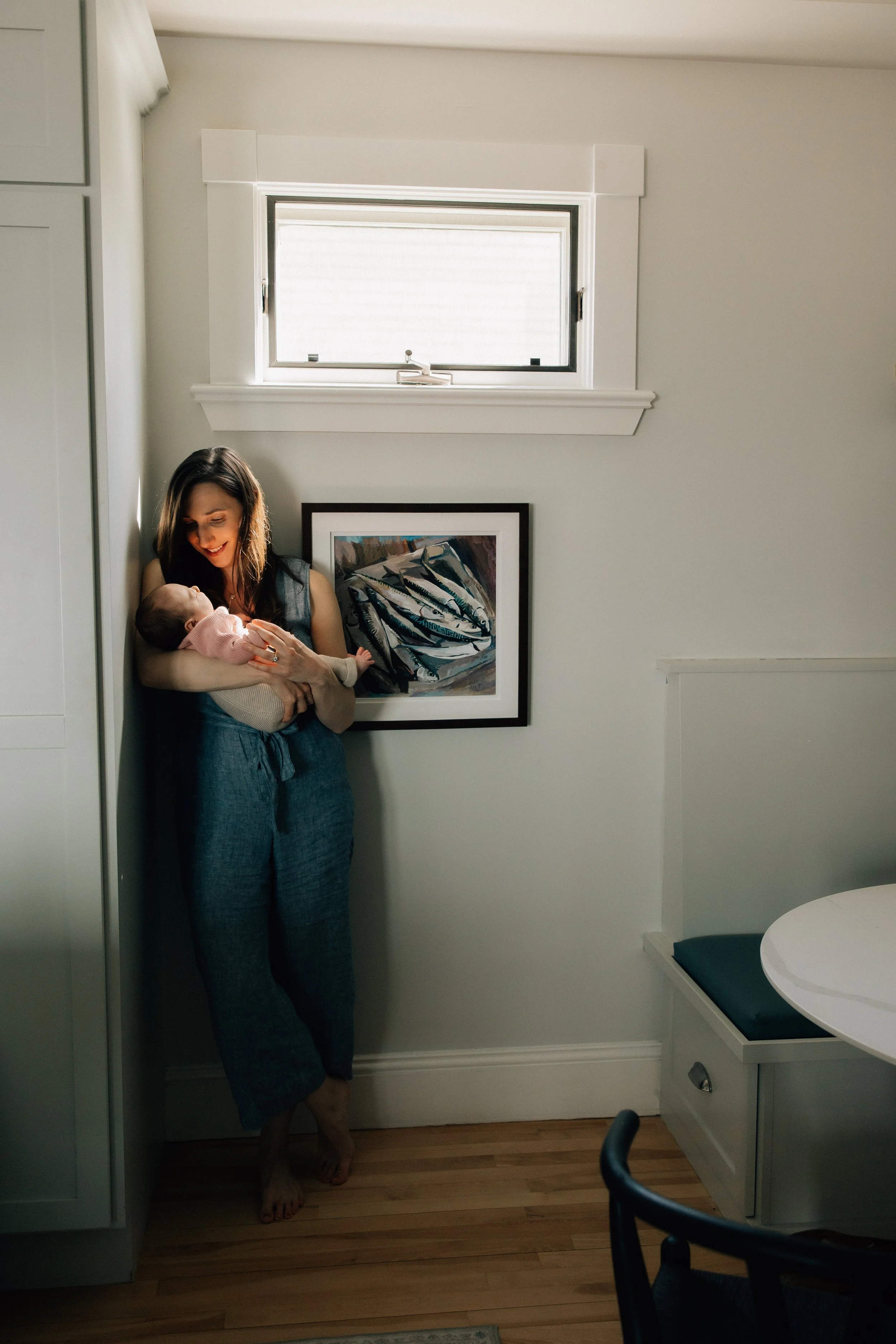 A woman holding a baby in a room with white walls, a window, artwork, and a small table with a blue cushion.