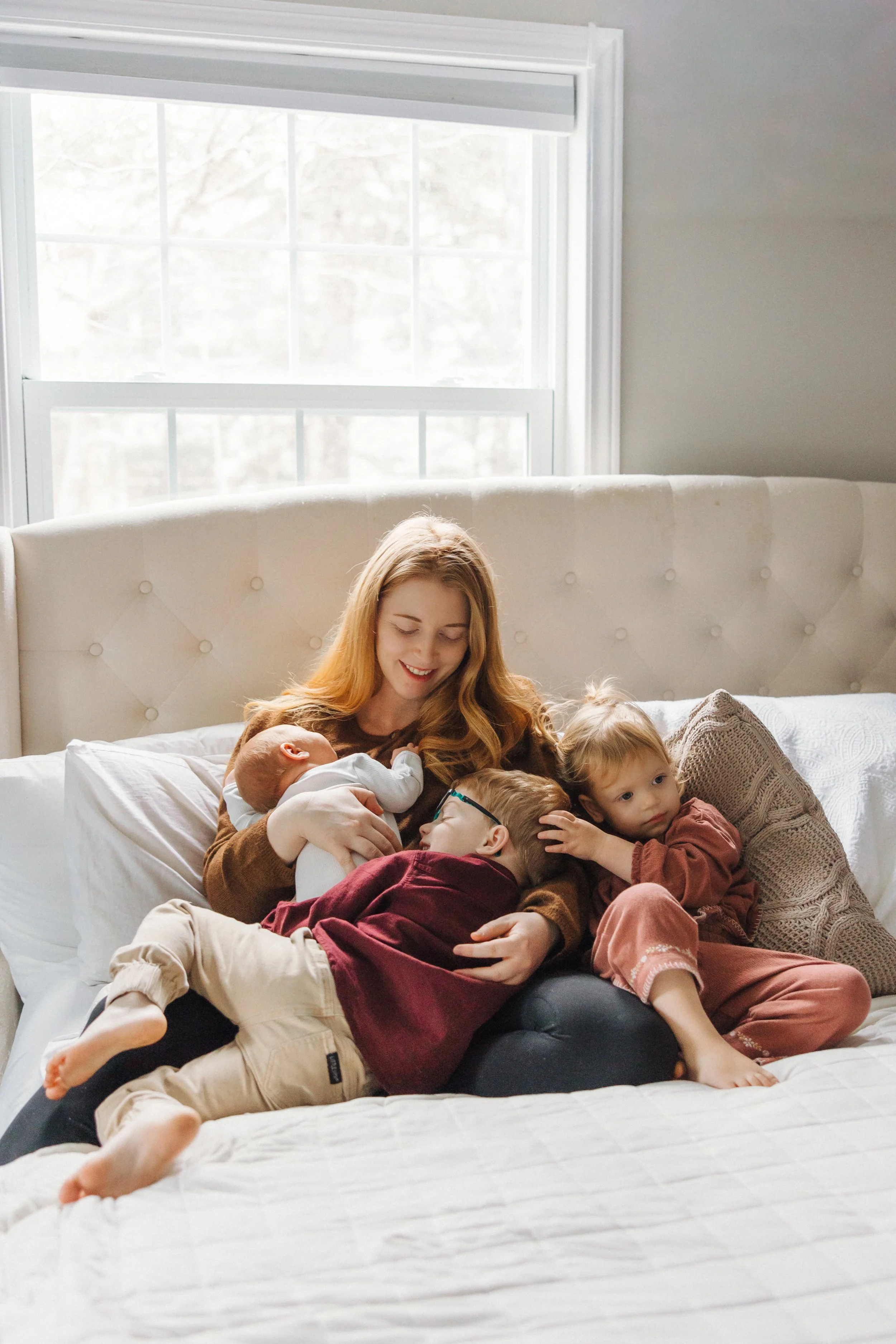 A woman with long red hair sitting on a bed with five children, all cuddling and smiling, in a bright room with a large window in the background.