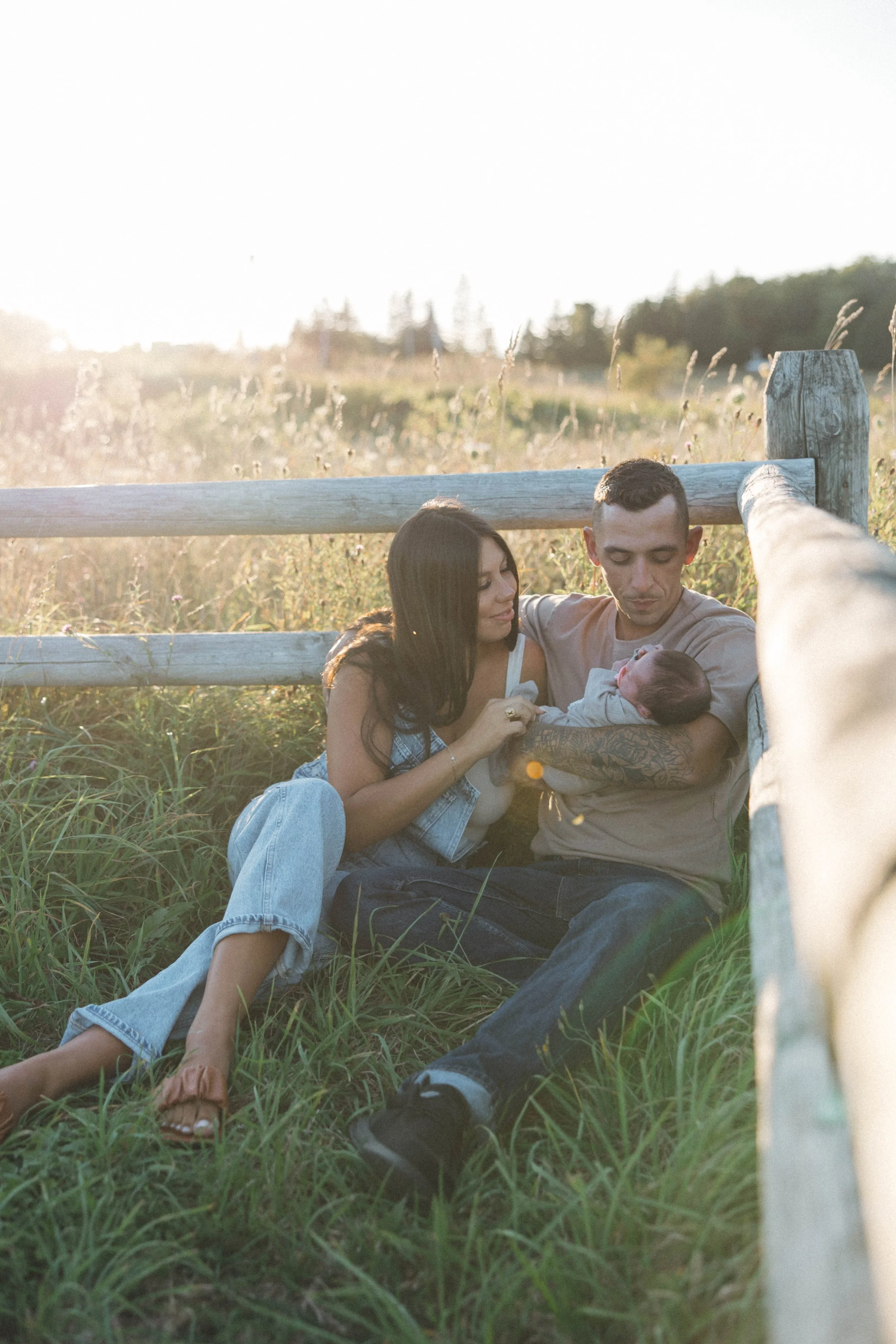 A family of four sitting on the grass by a wooden fence during sunset, with three adults, one holding a baby, and one a young girl, all smiling and enjoying the moment.
