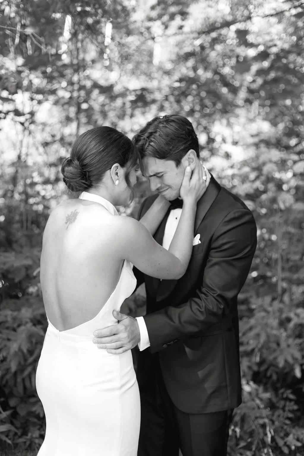 A black and white photo of a bride and groom embracing outdoors, with the bride holding the groom's face and the groom holding her waist, both smiling with eyes closed.