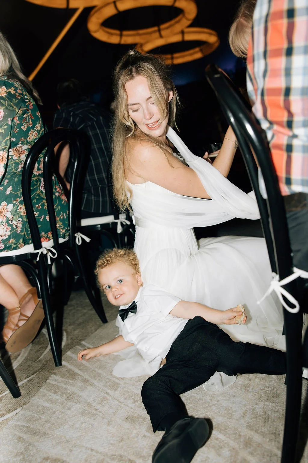 A woman in a white dress and a young boy in a white shirt with a bow tie sitting on the floor at a social event. The woman is smiling with her eyes closed, and the boy is lying on the carpet looking at the camera with a playful expression.