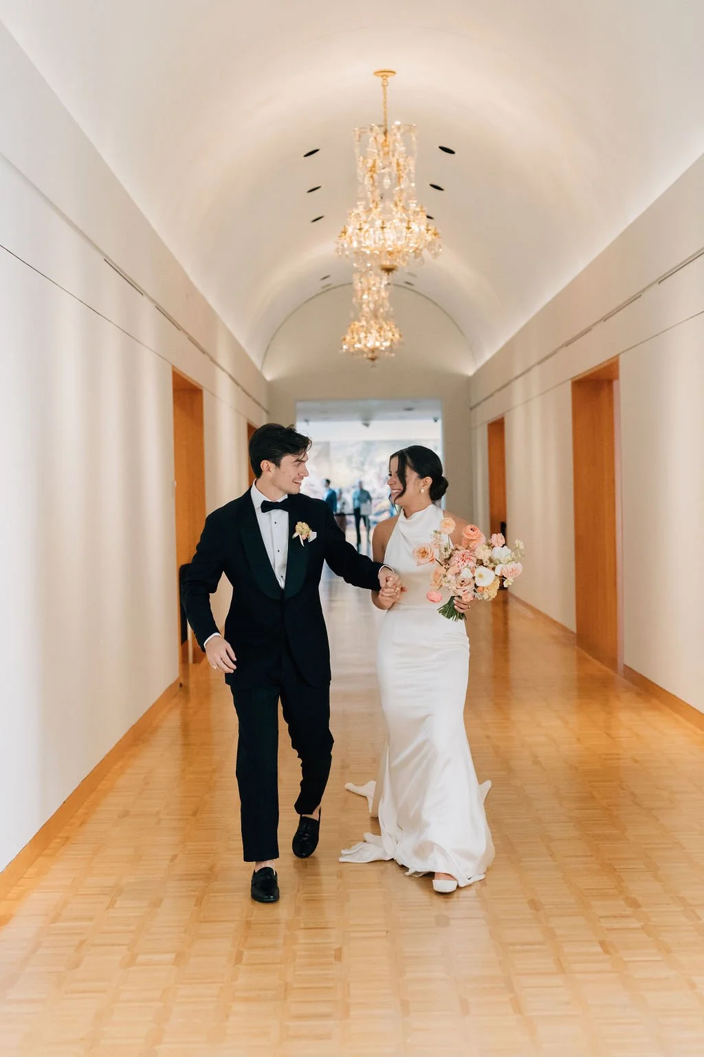 A bride and groom walking arm in arm down a hallway, smiling at each other. The bride is wearing a white wedding dress and holding a bouquet of pink and white flowers. The groom is wearing a black tuxedo with a bow tie. The hallway has a chandelier h