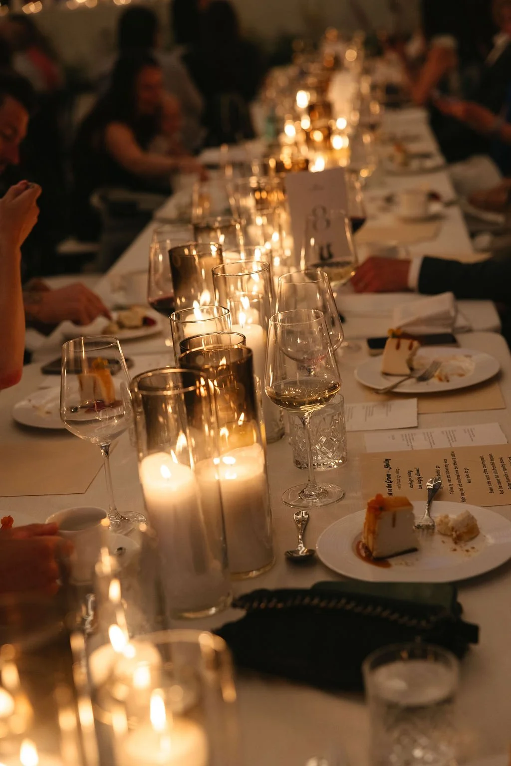 A long dinner table set for a formal event with lit candles in glass holders, empty wine glasses, plates with dessert, and place cards, with guests seated along both sides.